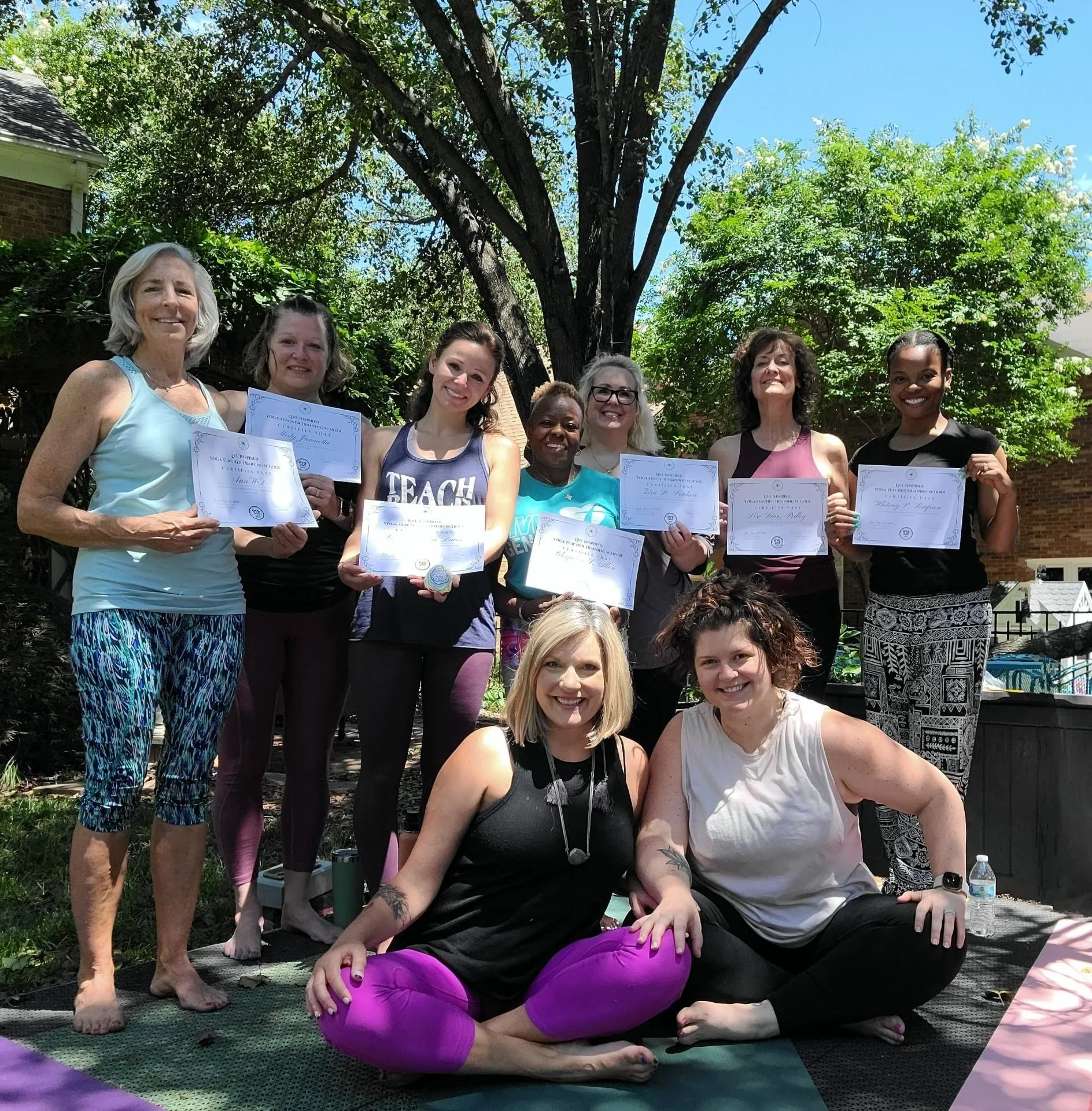 A group of women poses outdoors in front of a large tree and green foliage, holding certificates and smiling, with some seated on yoga mats and others standing behind them.