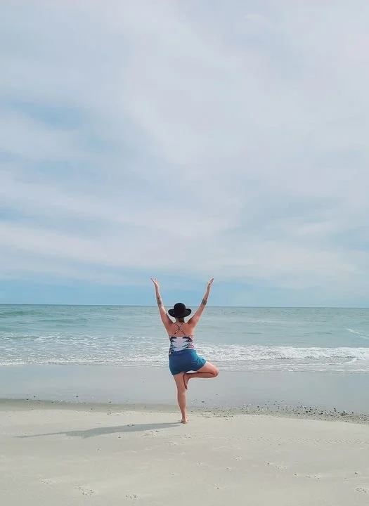 Woman practicing yoga on the beach facing the ocean with arms raised and one leg bent, wearing a black hat and summer clothes.