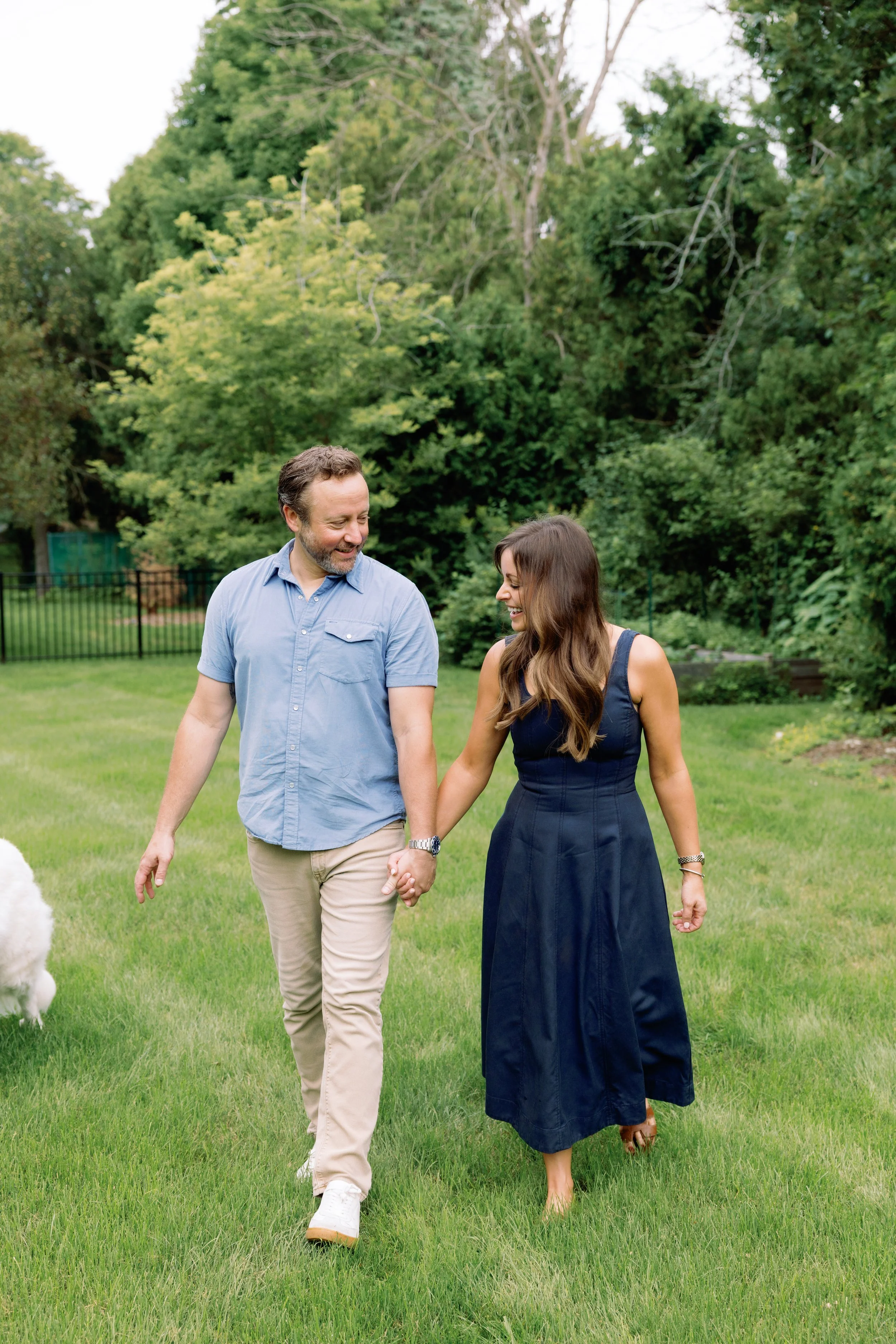 A man and woman walking hand-in-hand through a grassy park, smiling and looking at each other, with trees in the background.
