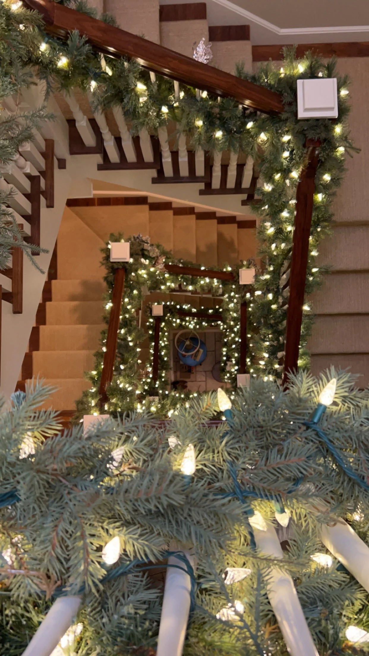 View of a spiral staircase decorated for Christmas with green garlands, white lights, and wrapped in holiday lights. The photo is taken from above by RWC, looking downward.