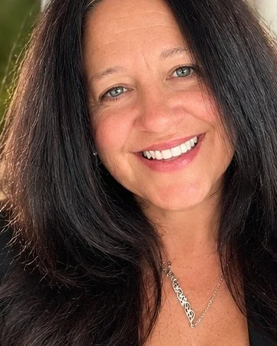 Close-up of a smiling woman with long dark hair, blue eyes, and wearing a necklace, outdoors.