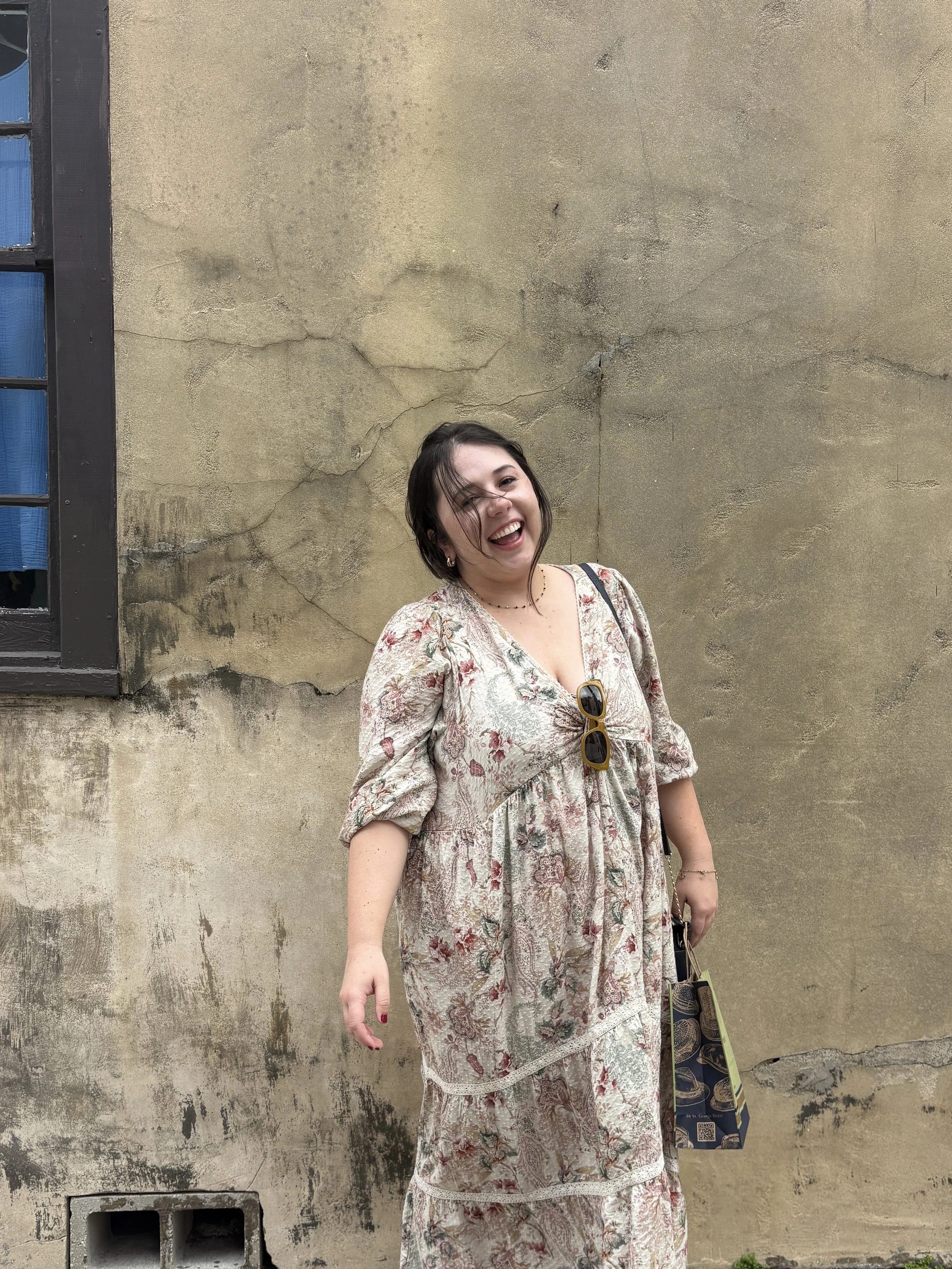 A smiling woman with short dark hair wearing a floral dress, holding a shopping bag, standing against a weathered beige wall.