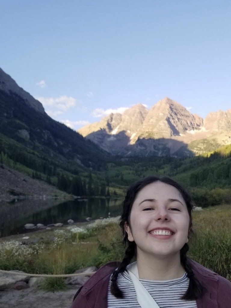 A young woman with braided hair smiling outdoors in front of a mountain landscape with a river, green hills, and snow-capped peaks in the background.