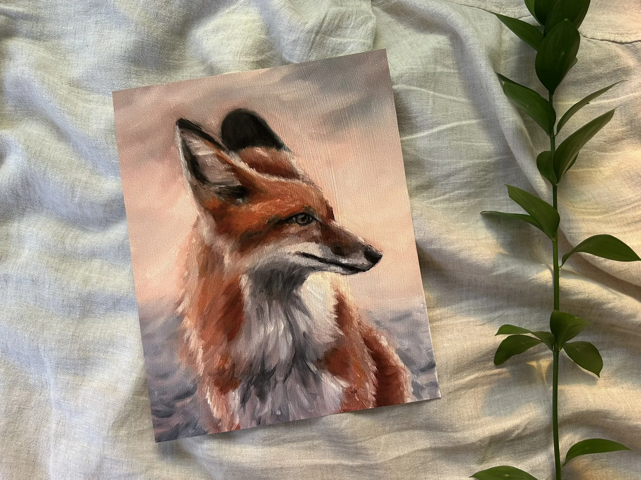 A watercolor painting of a fox with a focus on the face and upper body, depicting realistic fur details, placed on a soft, light-colored fabric with a green leafy plant next to it.