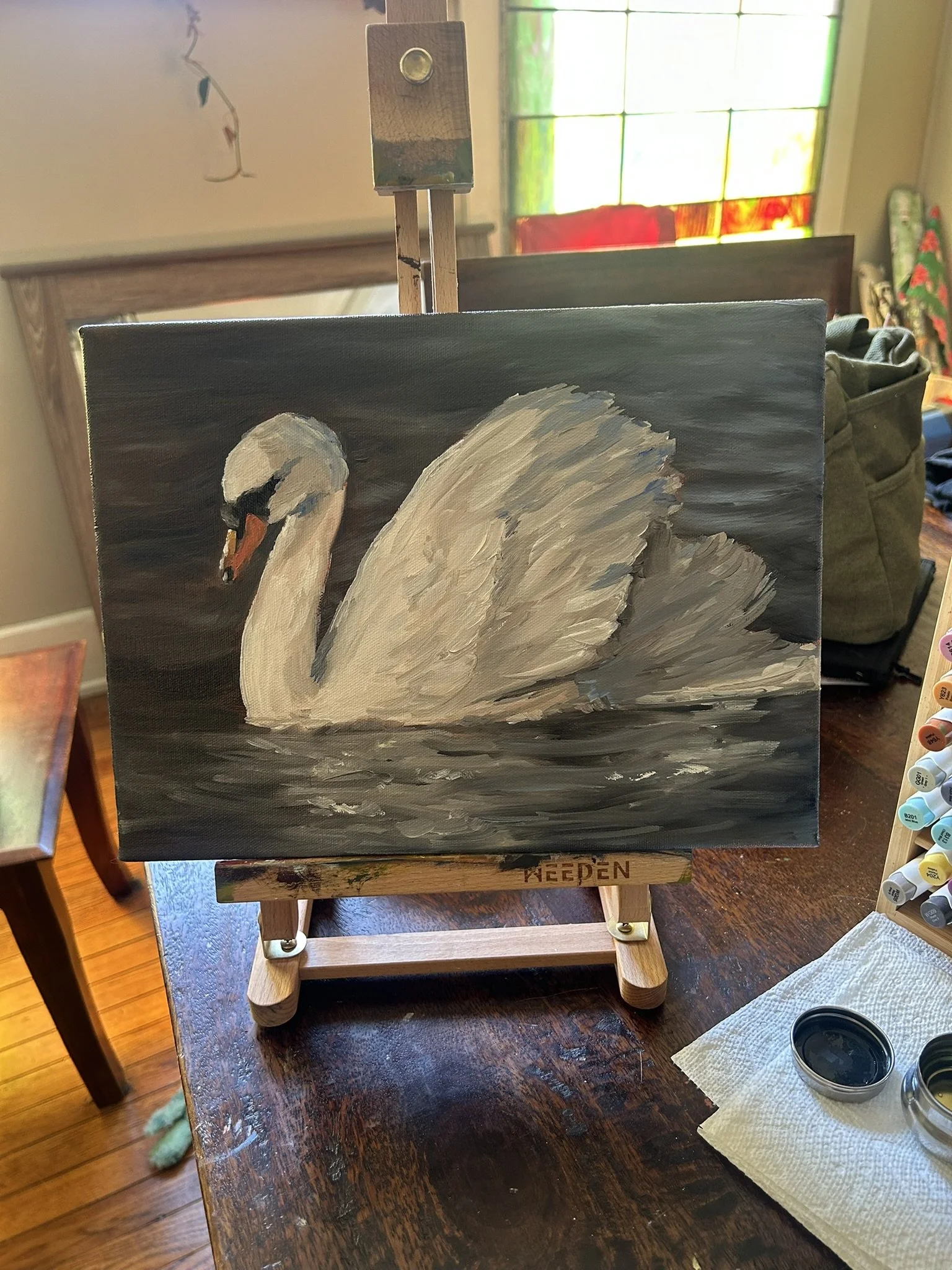 A painting of a swan is on an easel on a wooden table, with a container of black paint, a paper towel, and a palette to the right. In the background, there is a window with colorful stained glass.
