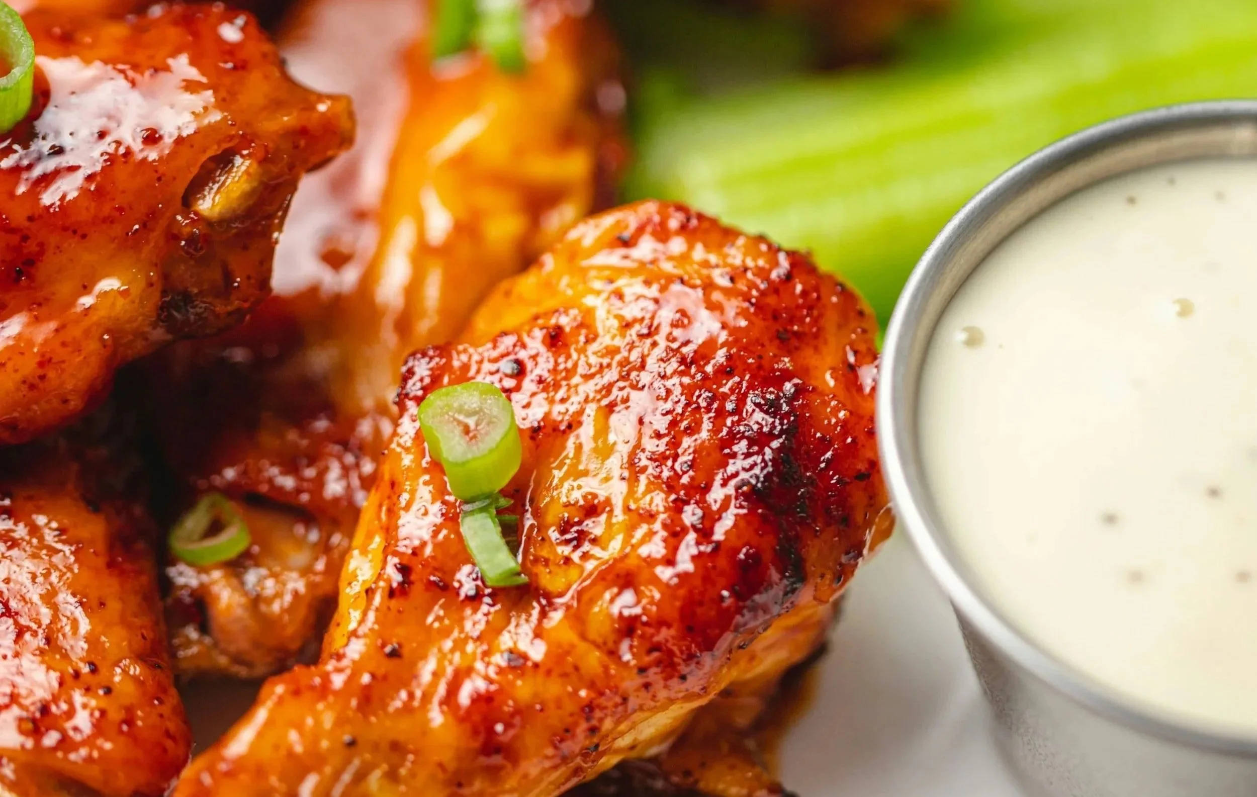 Close-up of glazed chicken wings garnished with chopped green onions, served with a side of creamy dipping sauce.