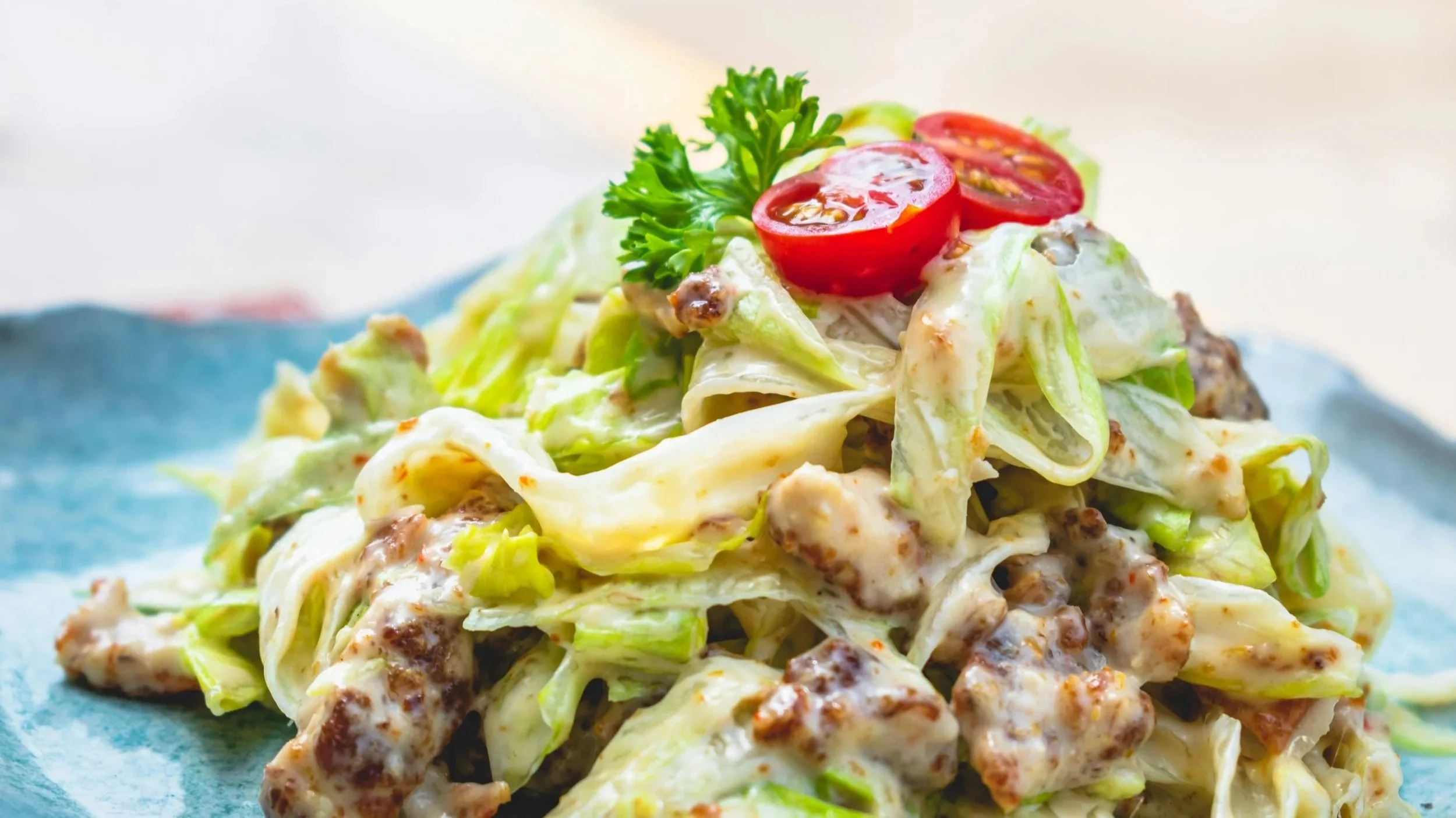 A close-up of a creamy chicken salad with chopped lettuce, cherry tomato halves, and fresh parsley served on a blue plate.