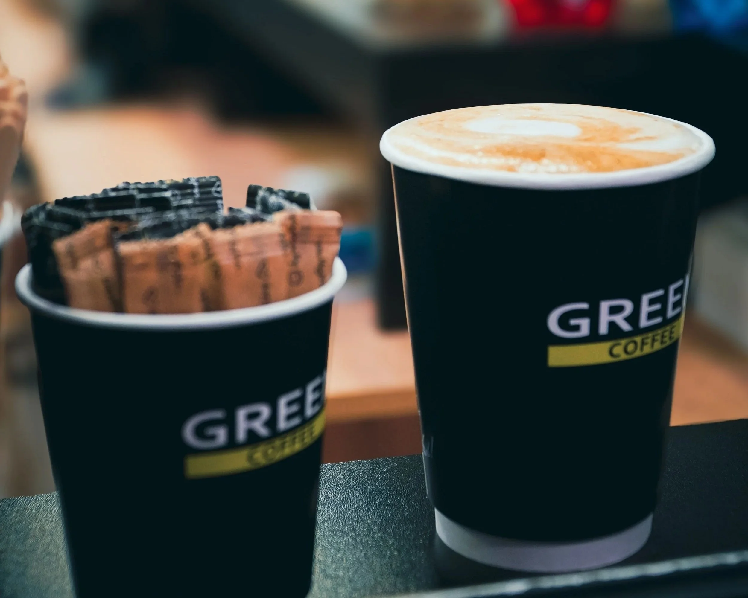 Two black paper cups filled with coffee, placed on a dark surface, with slight foam art visible on the coffee in each cup.