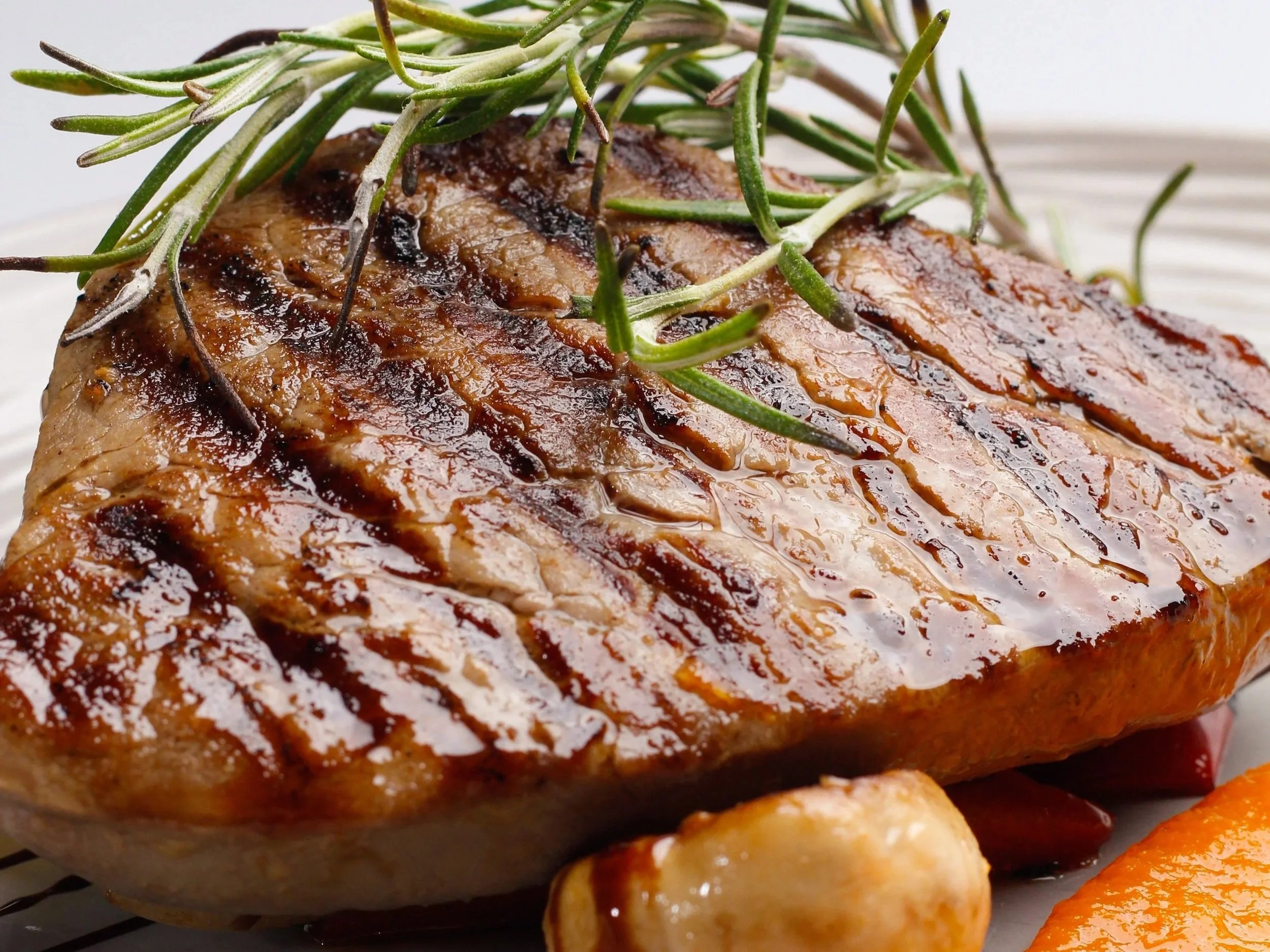 A close-up image of a cooked steak topped with rosemary sprigs, showing a glazed surface and grill marks.