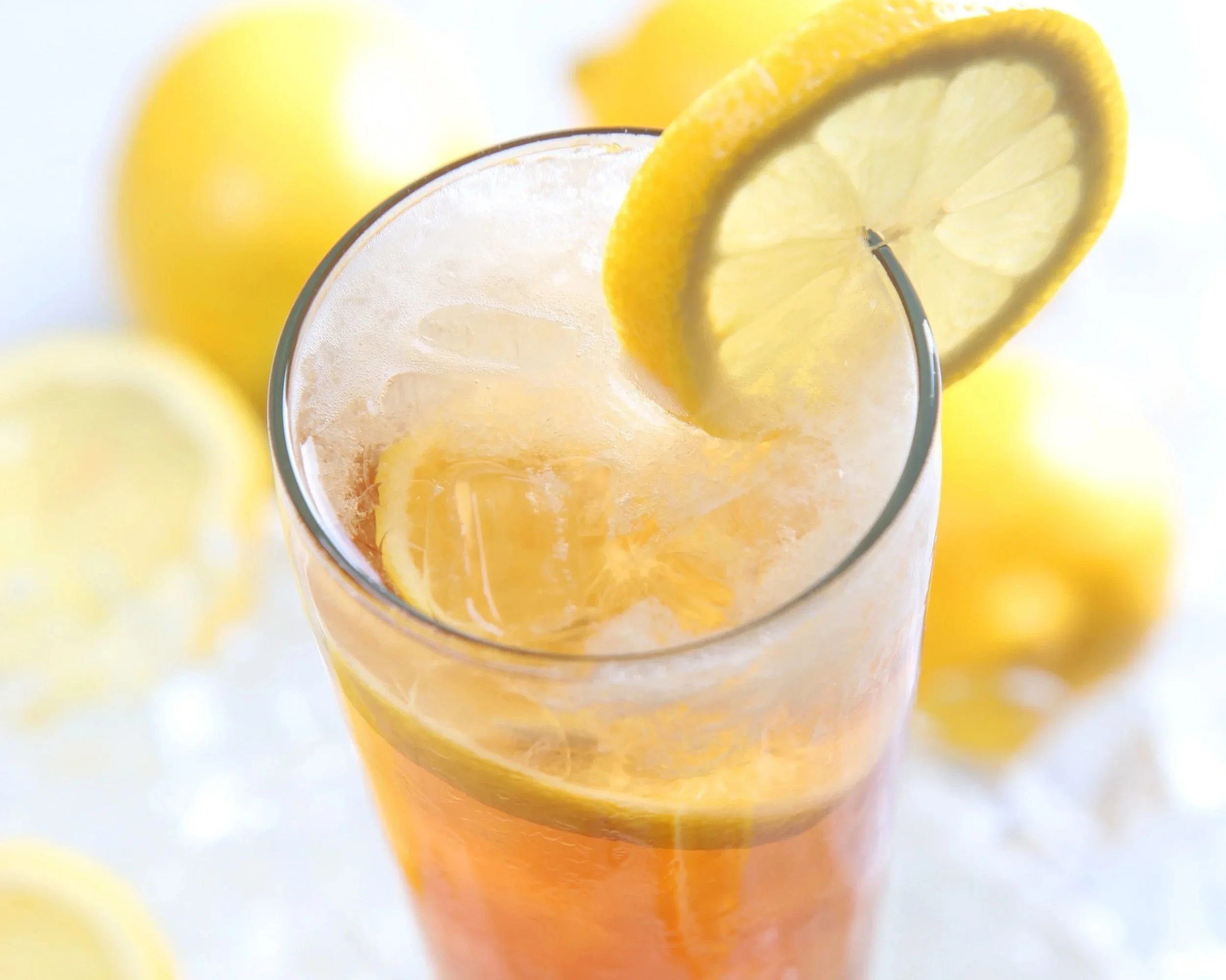 Close-up of a glass of lemonade with lemon slices, with blurred yellow lemons in the background.