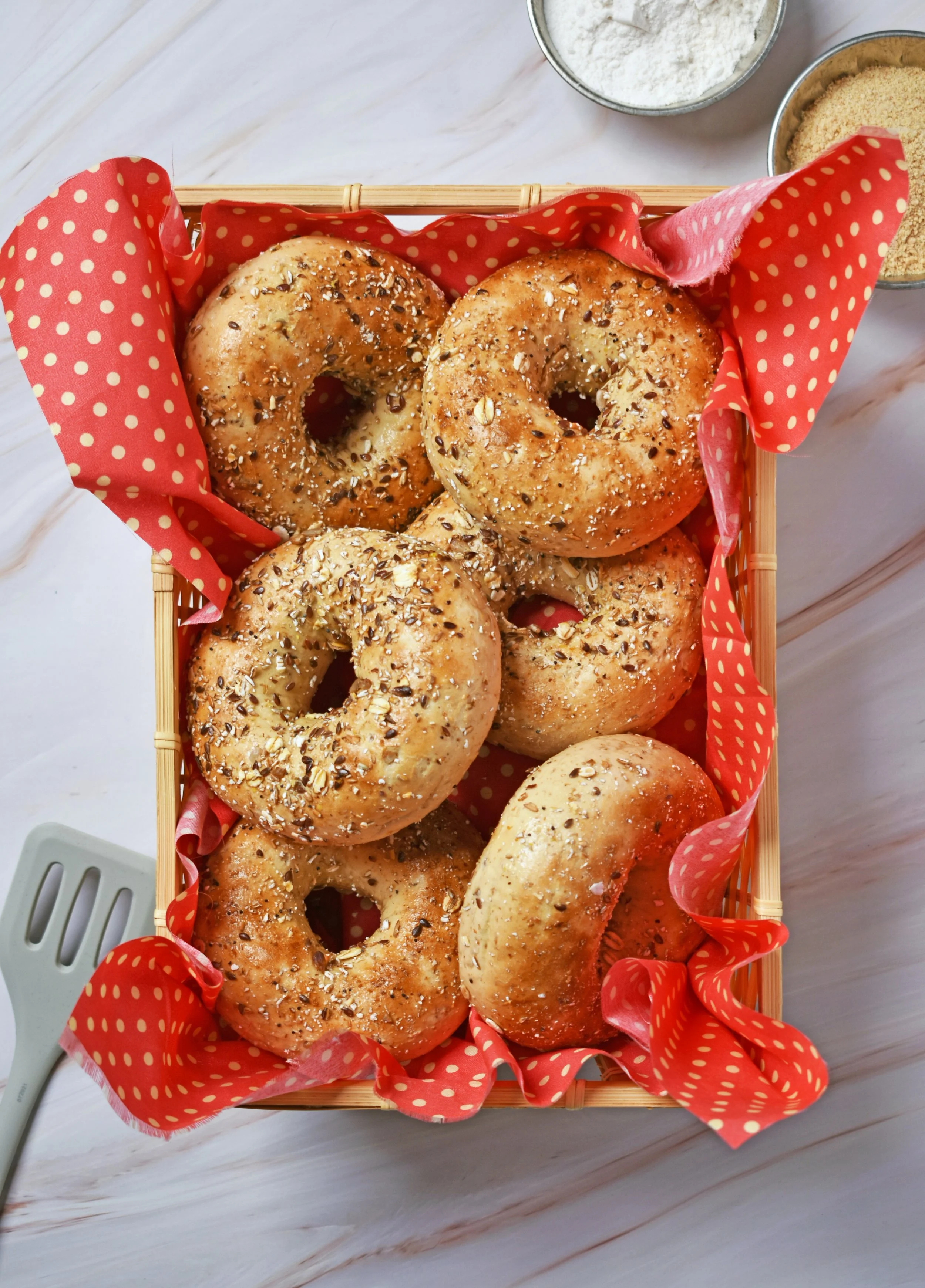 Basket of freshly baked bagels with various toppings in a red polka dot lining, placed on a white wooden surface with bowls of flour, sugar, and a spatula in the background.