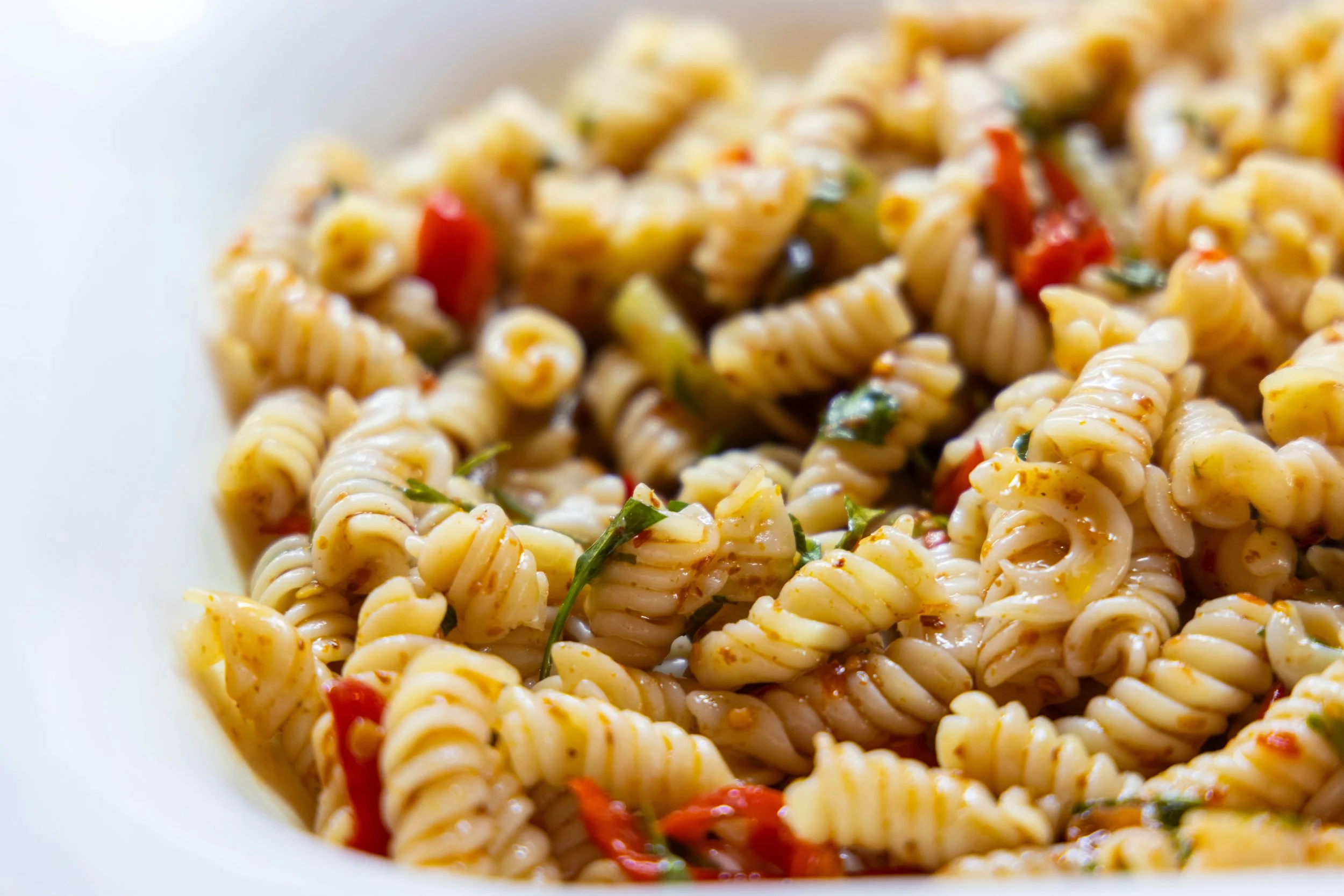Close-up of cooked fusilli pasta with red bell peppers, herbs, and sauce in a white bowl
