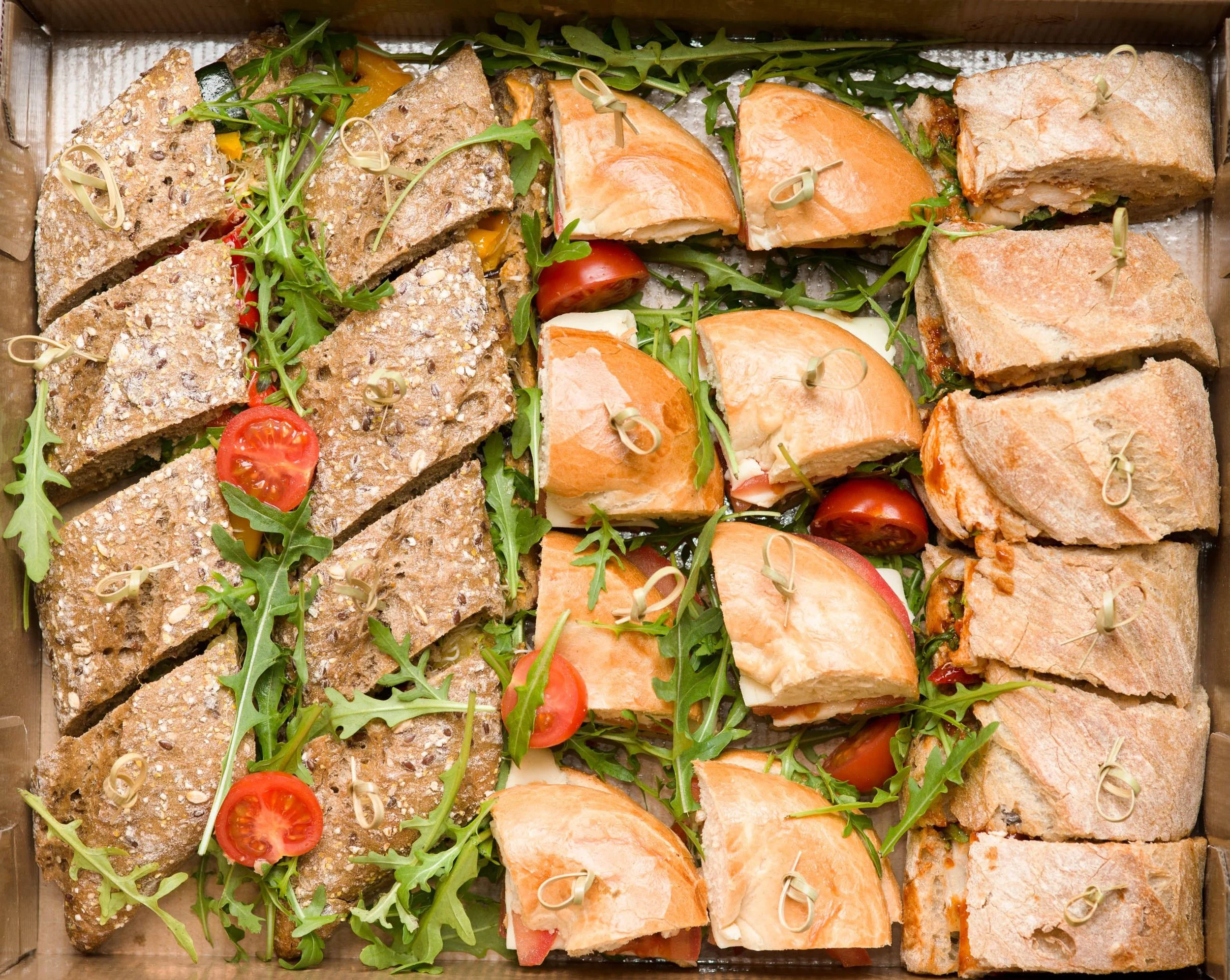 Assorted sandwiches with tomato, arugula, and slices of bread, topped with skewers, arranged on a tray.
