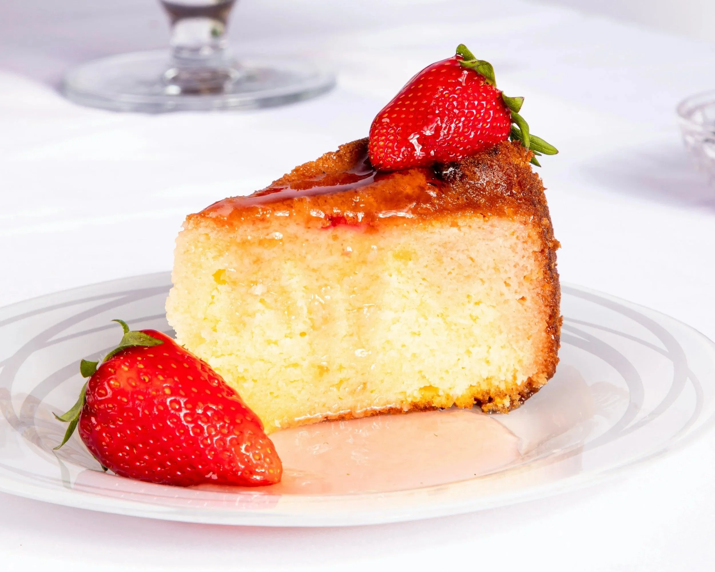 A slice of cheesecake topped with a whole strawberry and drizzled with strawberry syrup, on a white plate with additional strawberries nearby.