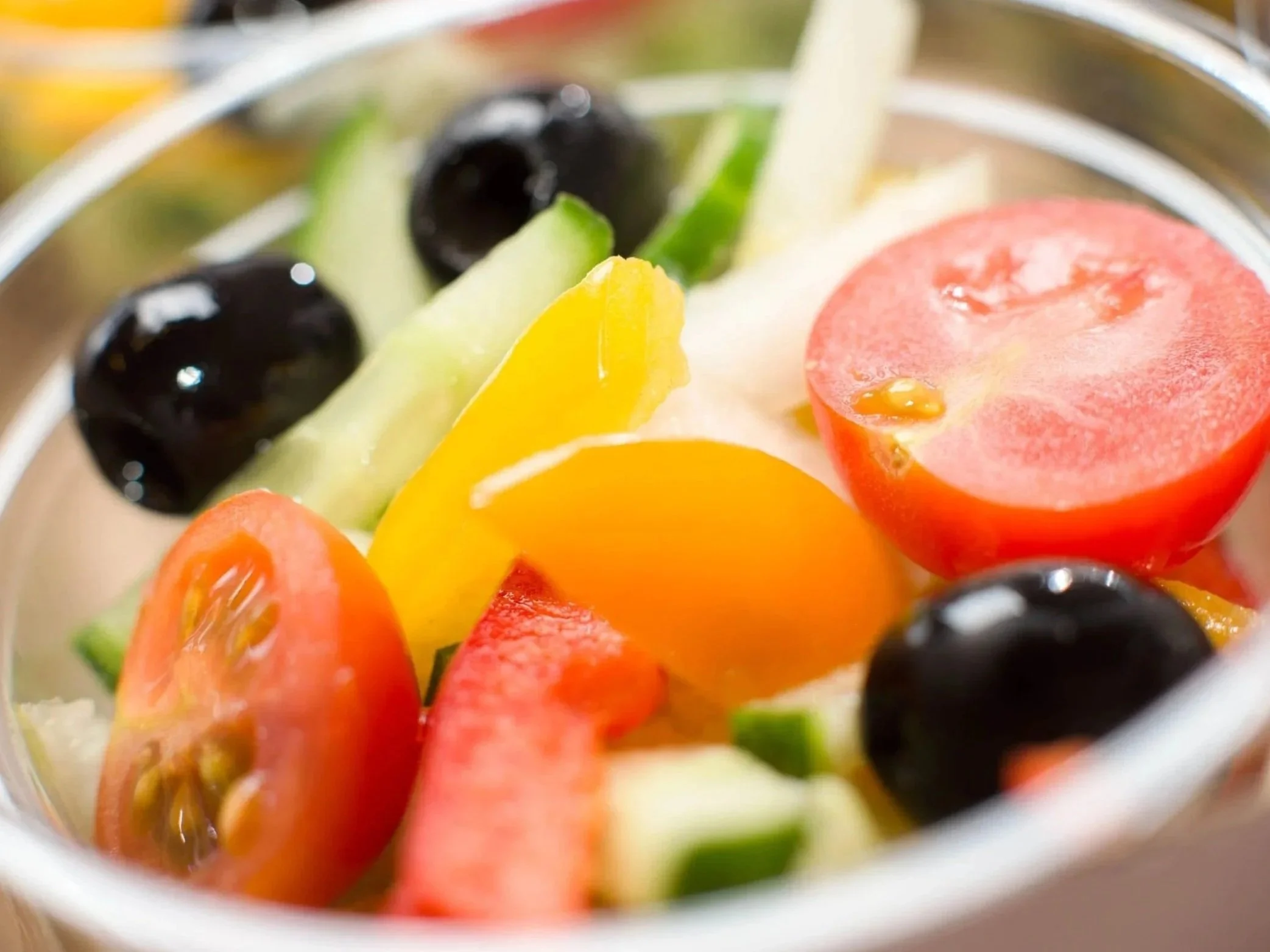 Close-up of fresh sliced tomatoes, cucumbers, black olives, and yellow bell peppers in a bowl.