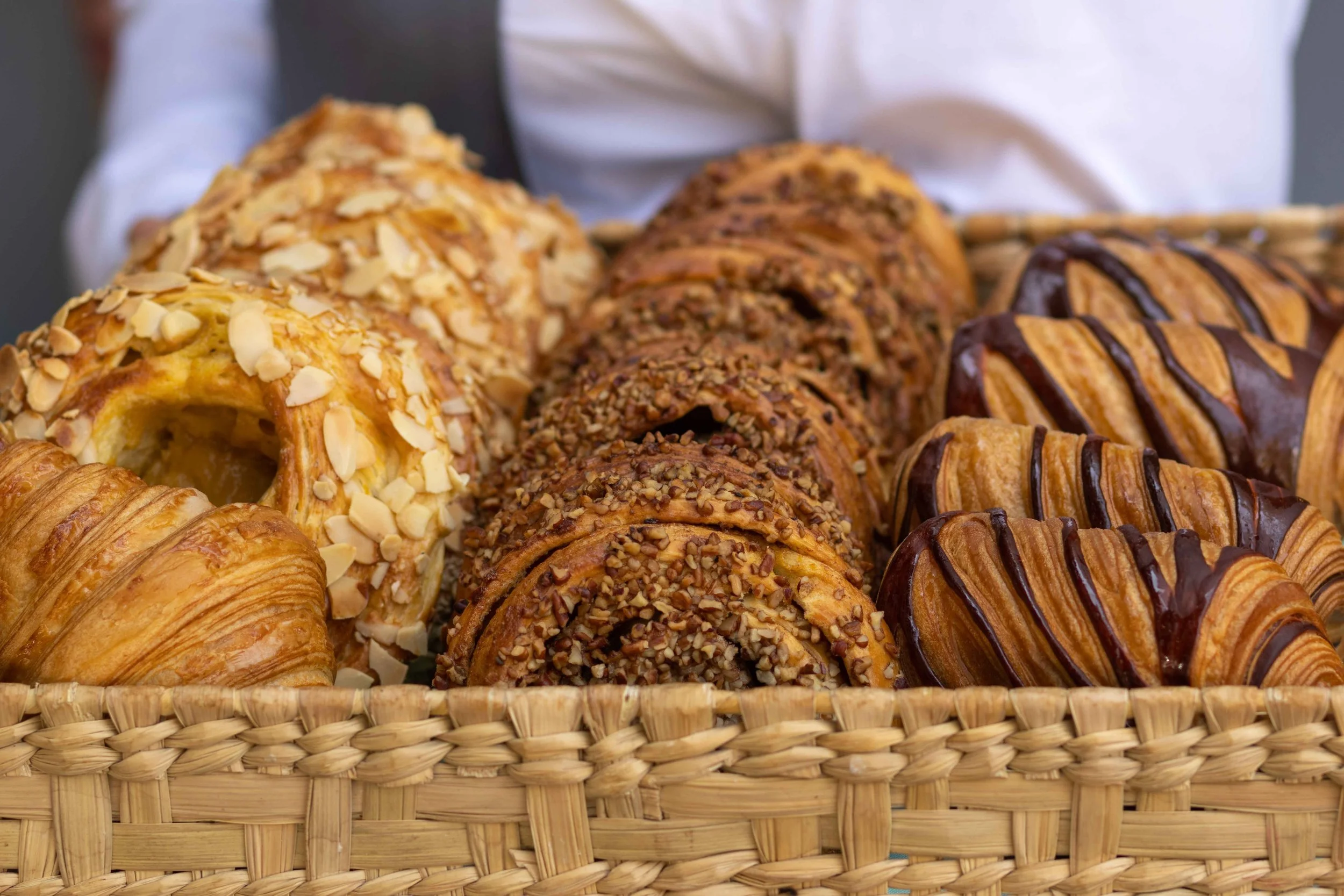A basket filled with assorted pastries, including croissants and Danish pastries topped with nuts and chocolate drizzle.