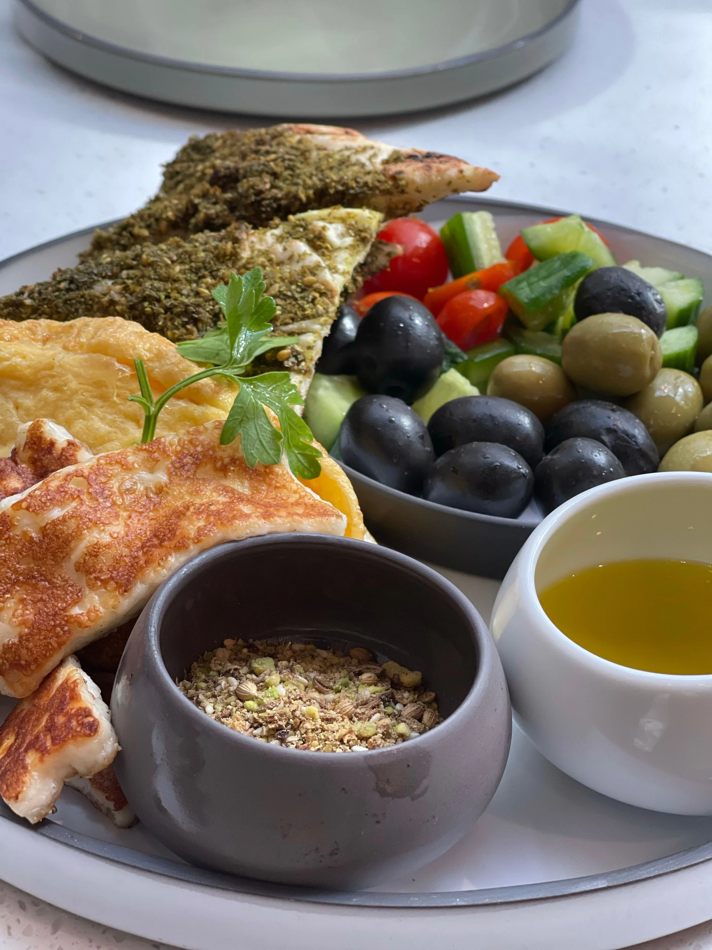 Close-up of a plate of food with a piece of fish coated in herbs, a slice of pizza, various vegetables like cherry tomatoes, cucumbers, green olives, black olives, and bowls of dips or sauces, on a white surface.
