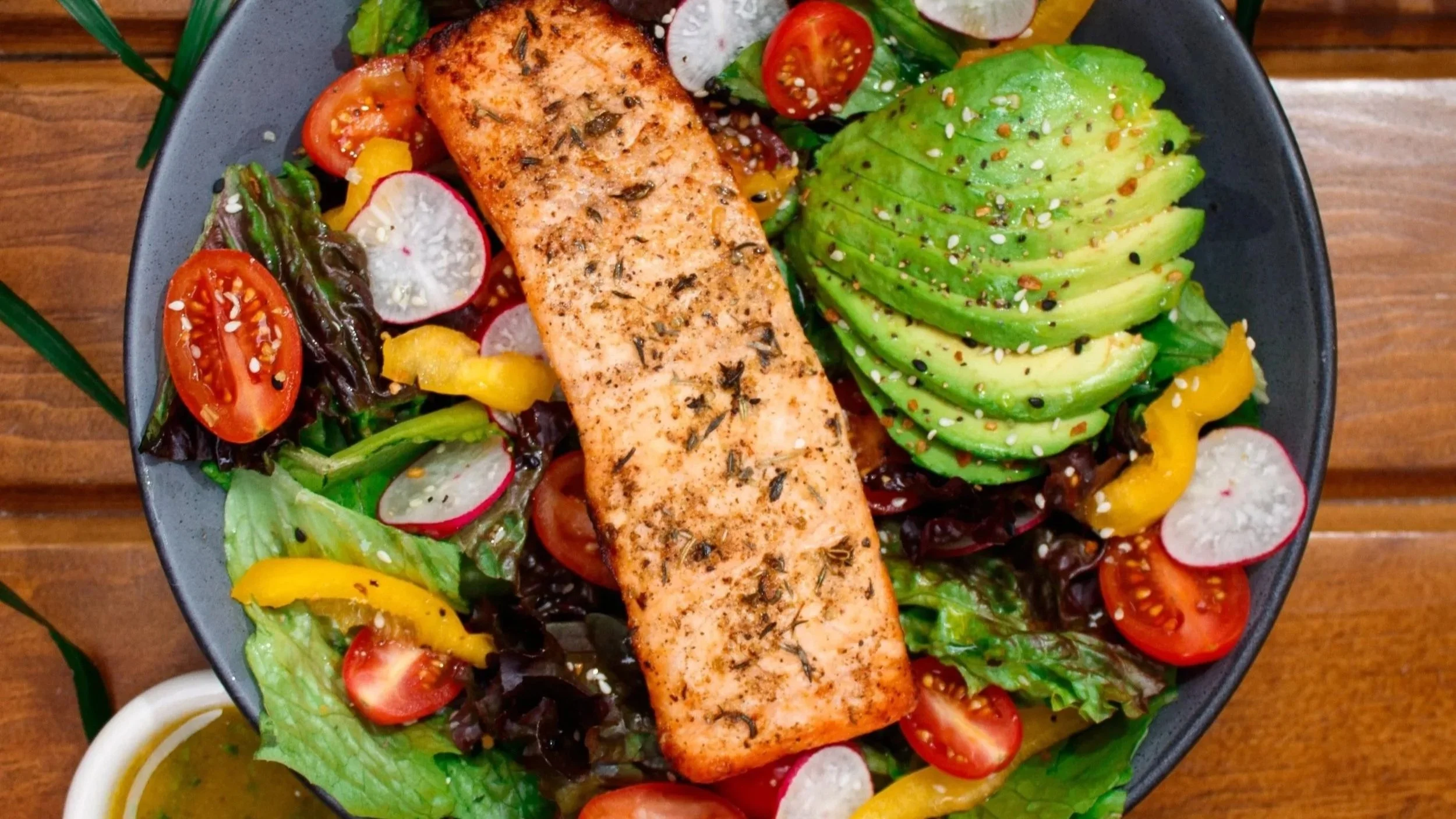 Salmon fillet on a bed of mixed greens, cherry tomatoes, radish slices, yellow peppers, and avocado slices, garnished with sesame seeds on a black plate.