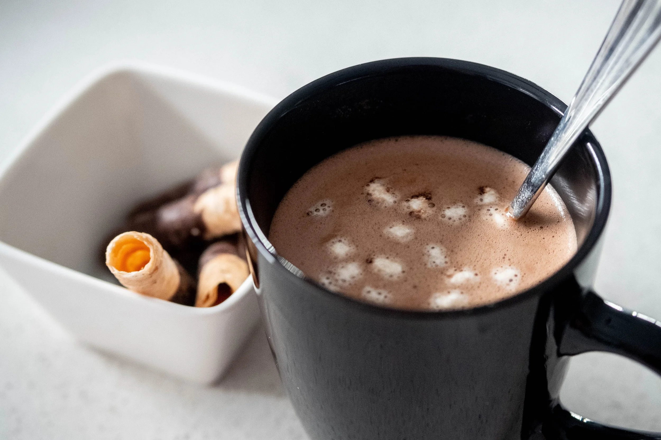 A close-up shot of a black mug filled with hot chocolate topped with marshmallows, with a spoon inside, placed on a white surface next to a white bowl containing rolled wafer cookies.