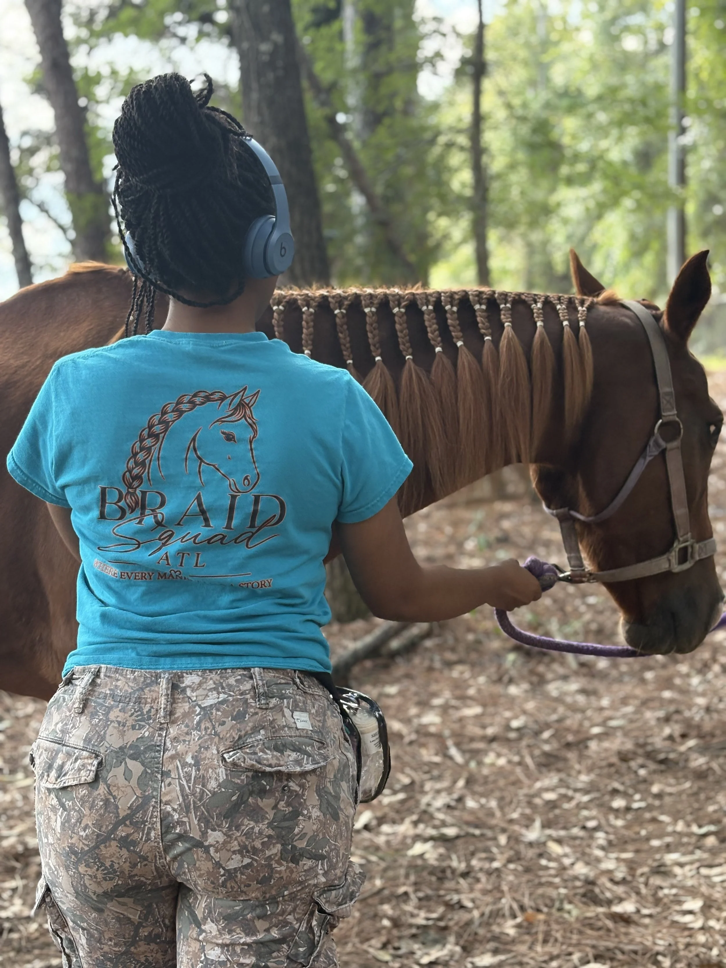 Tiarra (Tee Withers) braiding a horses mane, she’s wearing a blue T-shirt with a Braid Squad Atl logo and camouflage pants standing in a wooded area, holding the reins of a brown horse with a decorative braided mane.