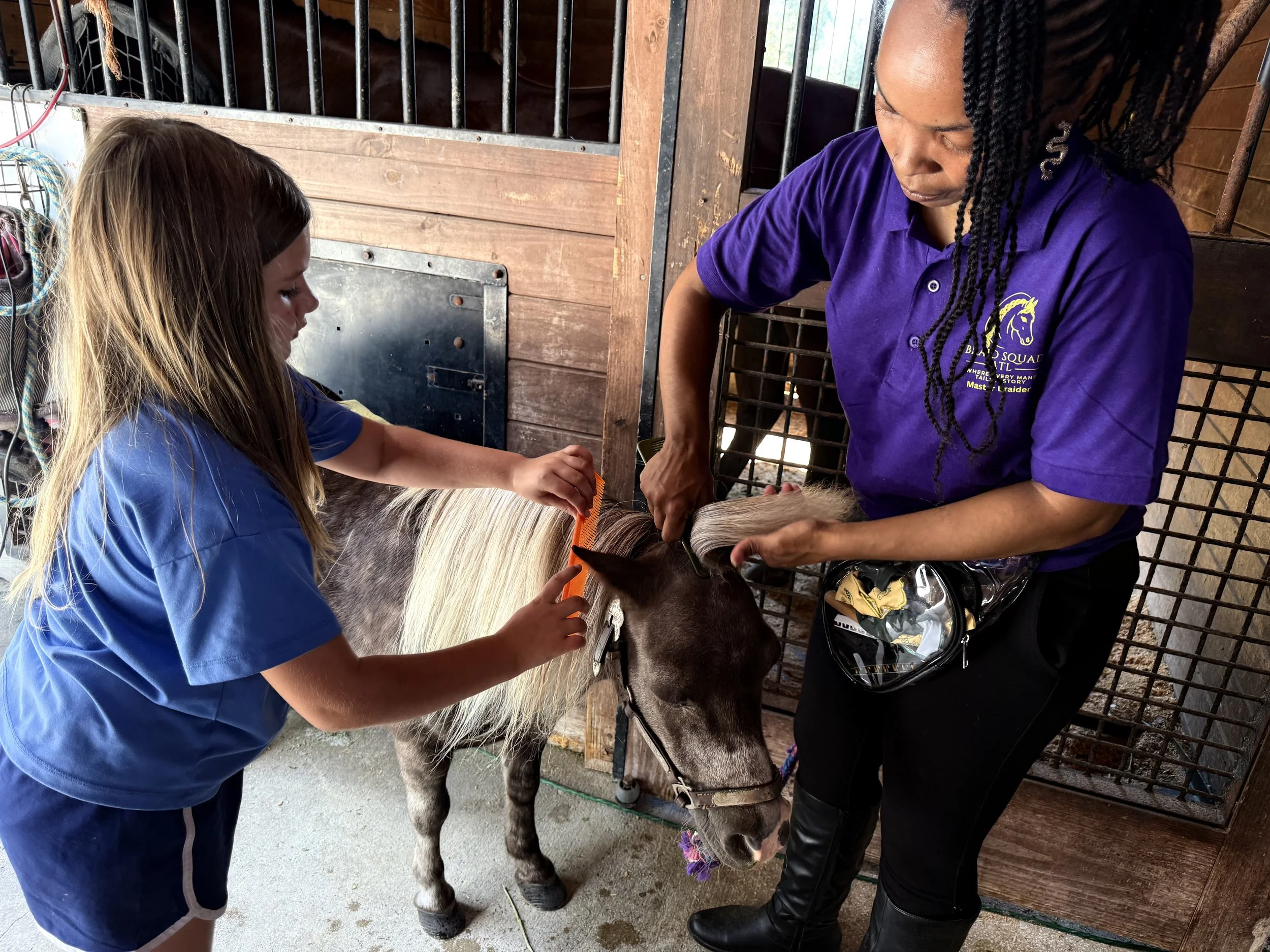 A young girl and a woman are tending to a small horse or pony inside a barn. The woman is teaching the girl to braid at the Braid Squad Atl hair clinic. The girl is brushing the horse's mane while the woman holds the horse's head gently.