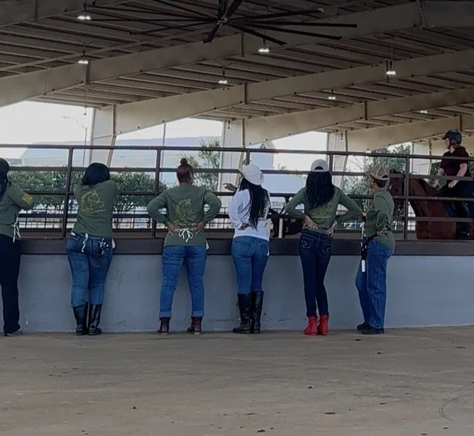 Six women at an indoor equestrian arena watching a horse being ridden by a person wearing a helmet. They are preparing to braid for a long night in the horse barn before show at fairgrounds in Perry Georgia.