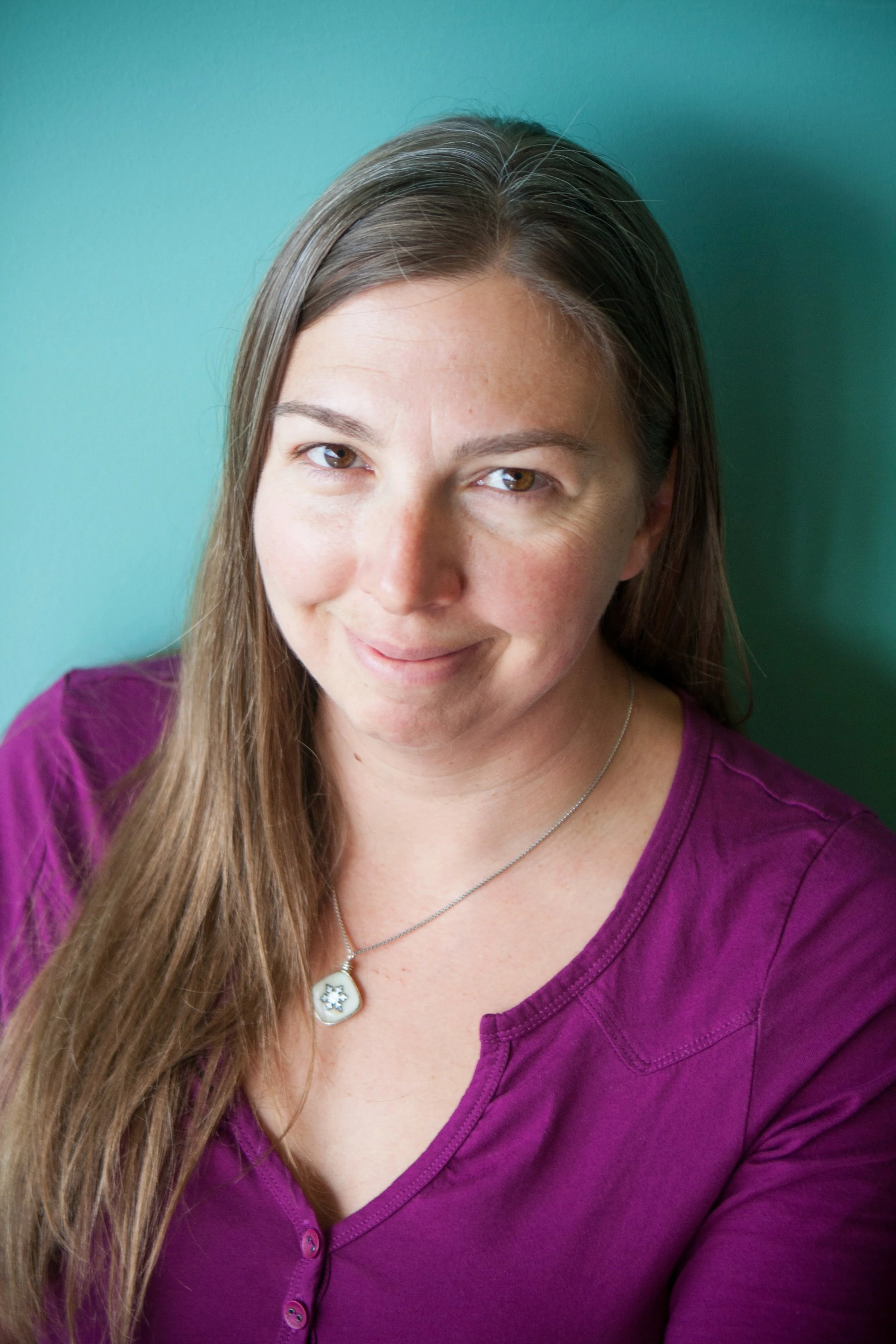 Close-up of a woman with long brown hair, wearing a purple shirt and a necklace with a snowflake pendant, smiling slightly in front of a teal background.
