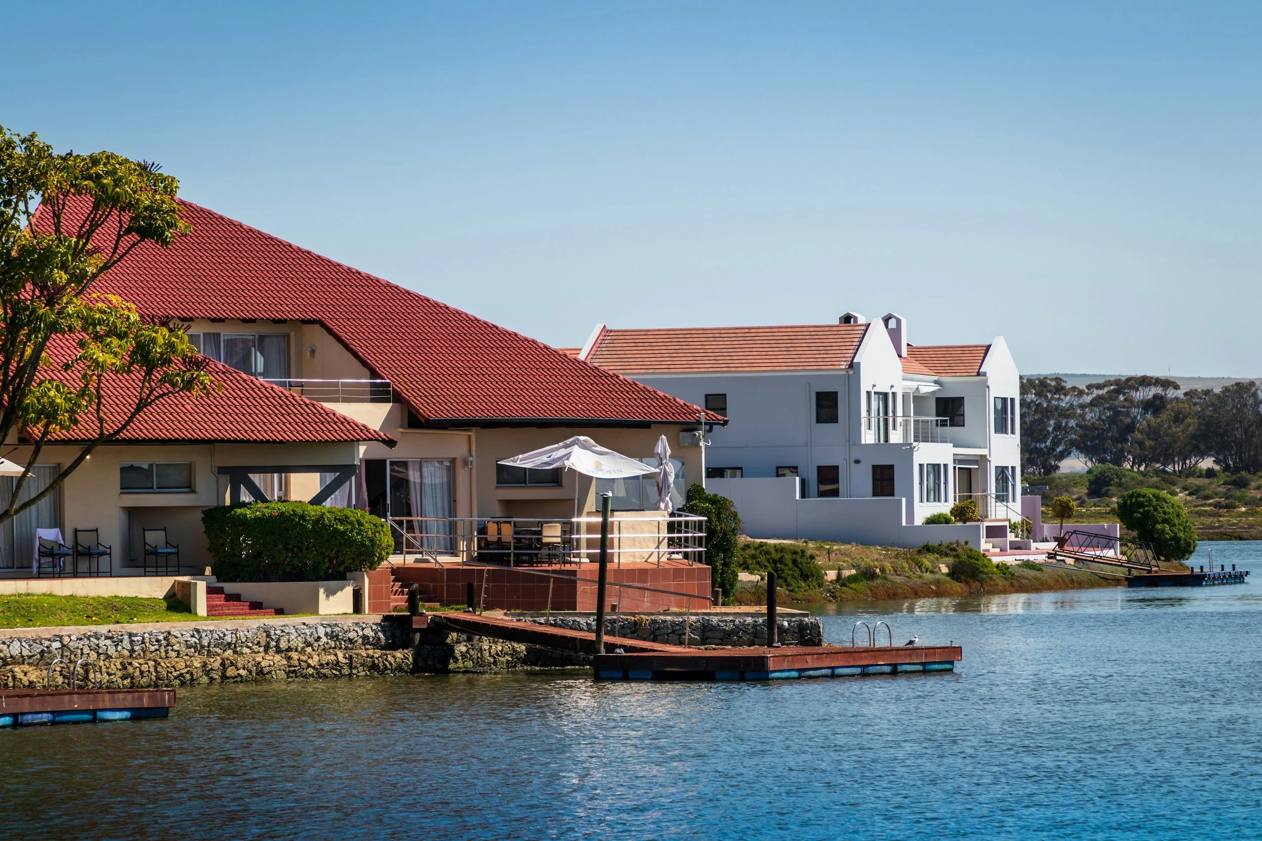 A lakeside scene with residential houses featuring red and white rooftops, a tree in the foreground, a dock extending into the water, and clear blue skies.