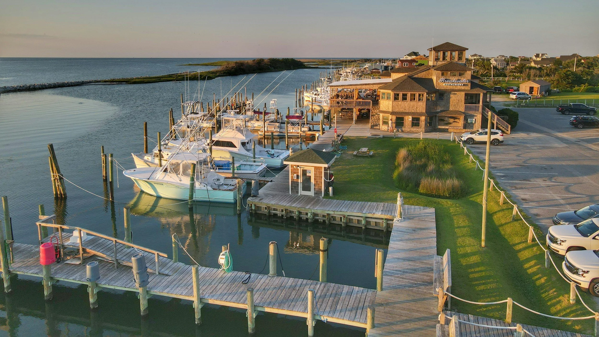 A marina with boats docked along wooden piers, a restaurant called 'Breakwater,' and a parking lot filled with cars. The scene is set during late afternoon or early evening with calm water and a clear sky.
