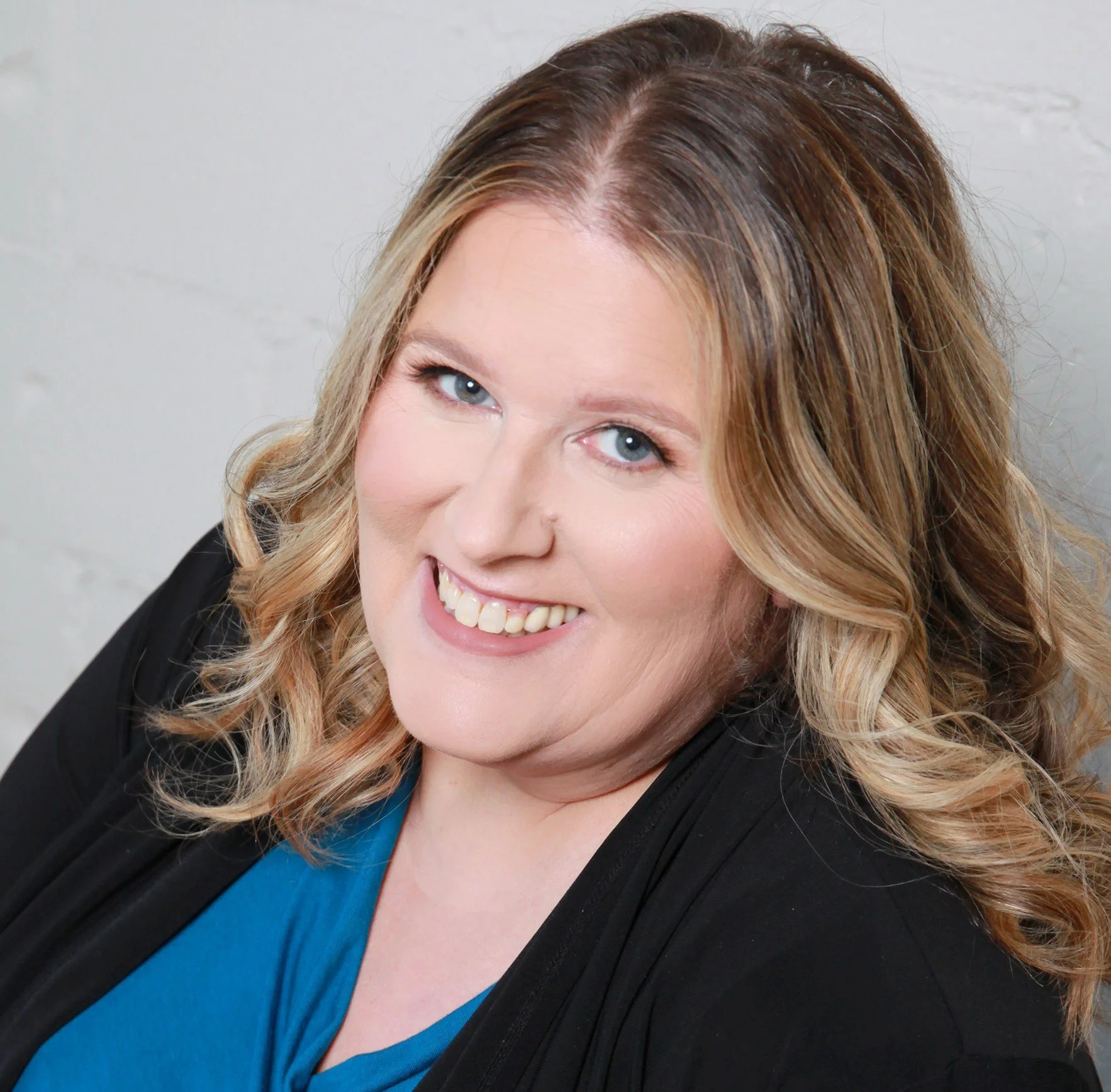 Close-up portrait of a woman with blonde, wavy hair, blue eyes, smiling, wearing a black blazer and blue top, against a white brick wall background.