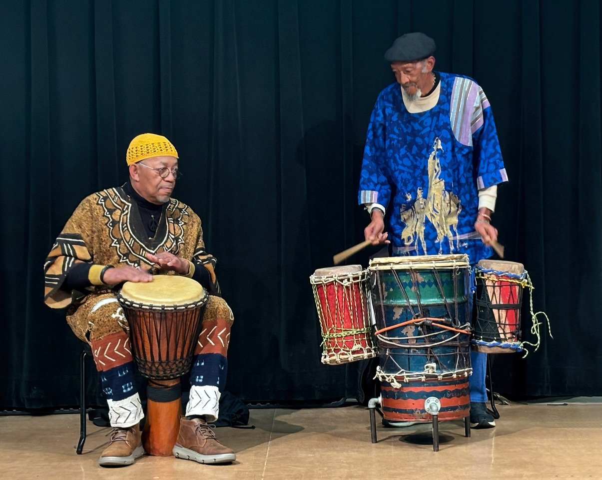 Two men playing drums on stage, one seated and wearing colorful traditional African clothing with a yellow hat, and the other standing and wearing a blue outfit with a black cap, against a black curtain background.