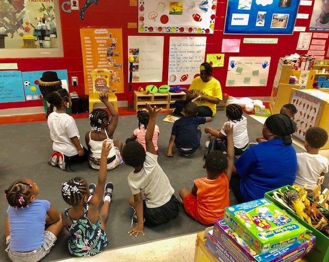 A teacher reading a book to a group of children sitting on the floor in a preschool classroom. Several children have their hands raised, indicating they want to answer or ask a question. The classroom has colorful walls with educational posters and t
