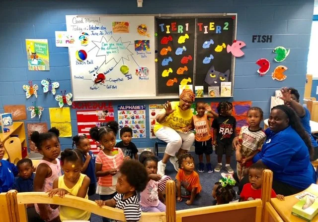 A group of young children in a colorful classroom with teachers, surrounded by educational posters and decorations, including butterflies, fish, and a whiteboard with notes.