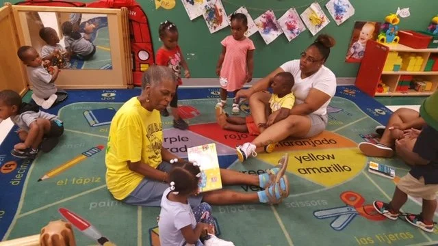 Adults and children sitting on a colorful educational rug in a classroom, with books, toys, and artwork on the walls.