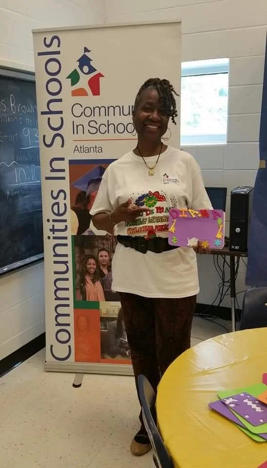 A woman smiling and holding a colorful decorated card and a box, standing next to a banner that reads 'Communities In Schools Atlanta.'