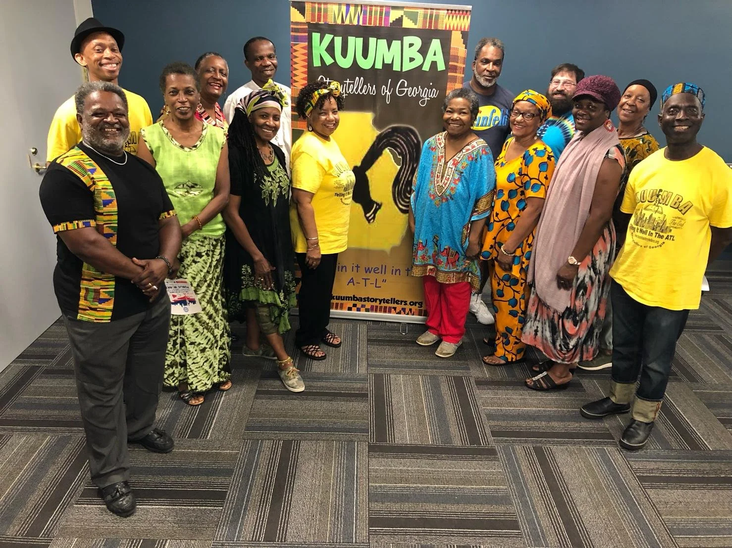 Group of diverse people standing in front of a colorful banner that reads "KUUMBA Storytellers of Georgia." They are dressed in traditional and casual clothing, smiling at the camera.