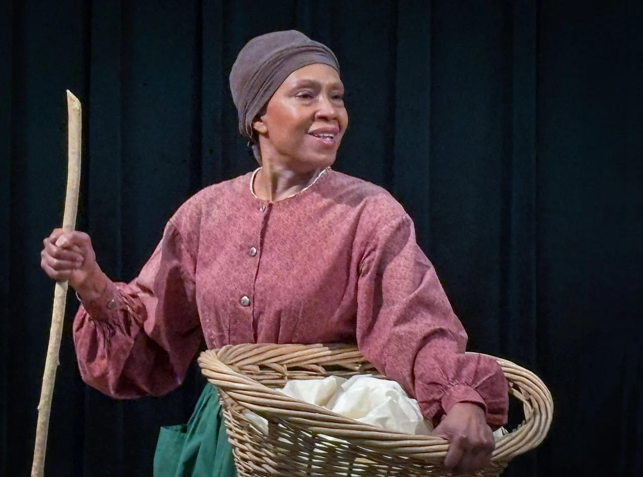 Traditional African woman wearing a headwrap and holding a walking stick, standing with a basket on her waist against a black background.