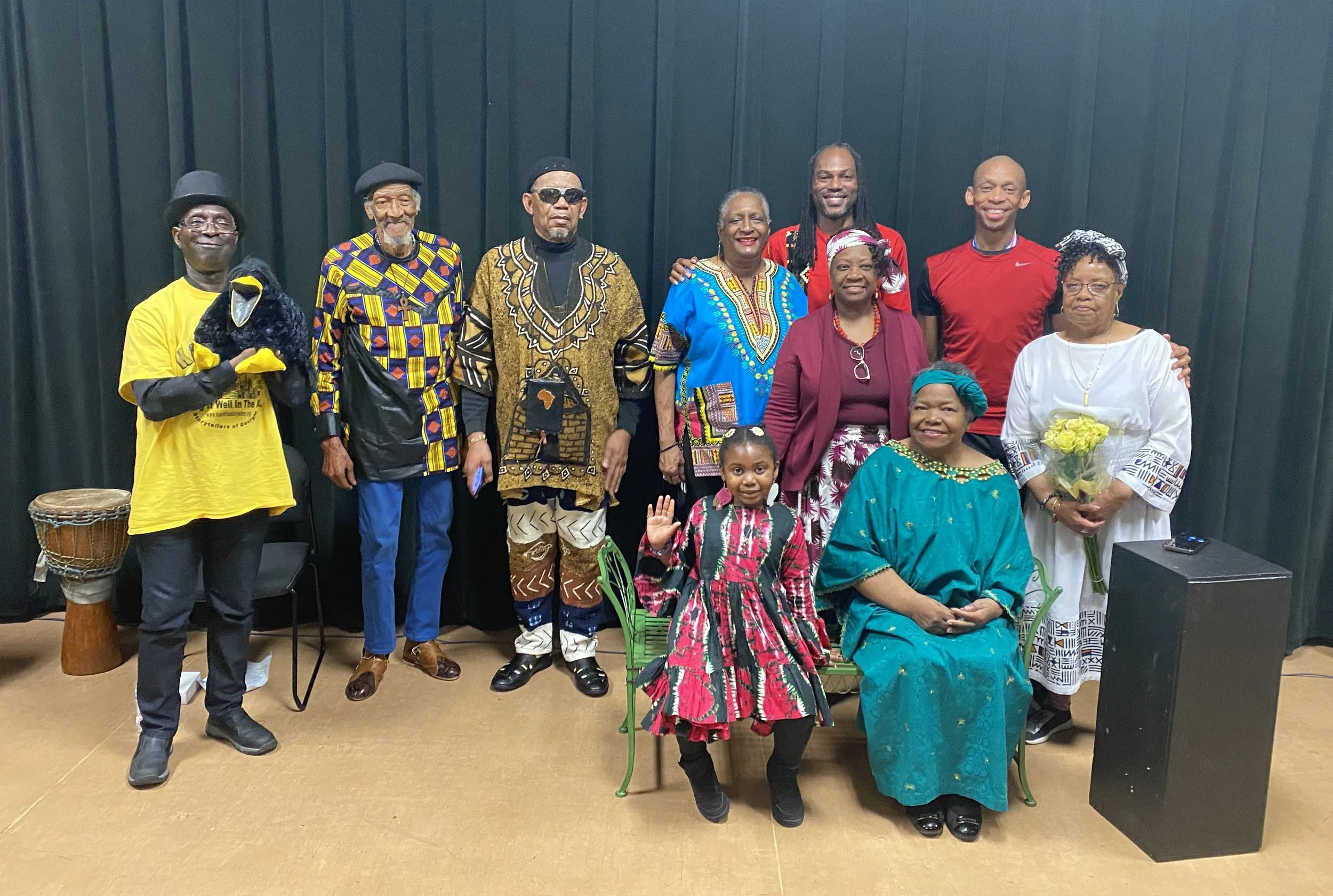 A group of diverse people posing together indoors, some wearing traditional African attire and others casual clothing, with a black curtain background.