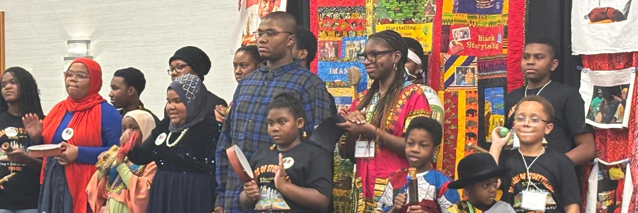 A group of children and adults standing on a stage, some holding items like books, percussion instruments, and maracas, in front of a colorful quilt display with various African-inspired patterns and images.
