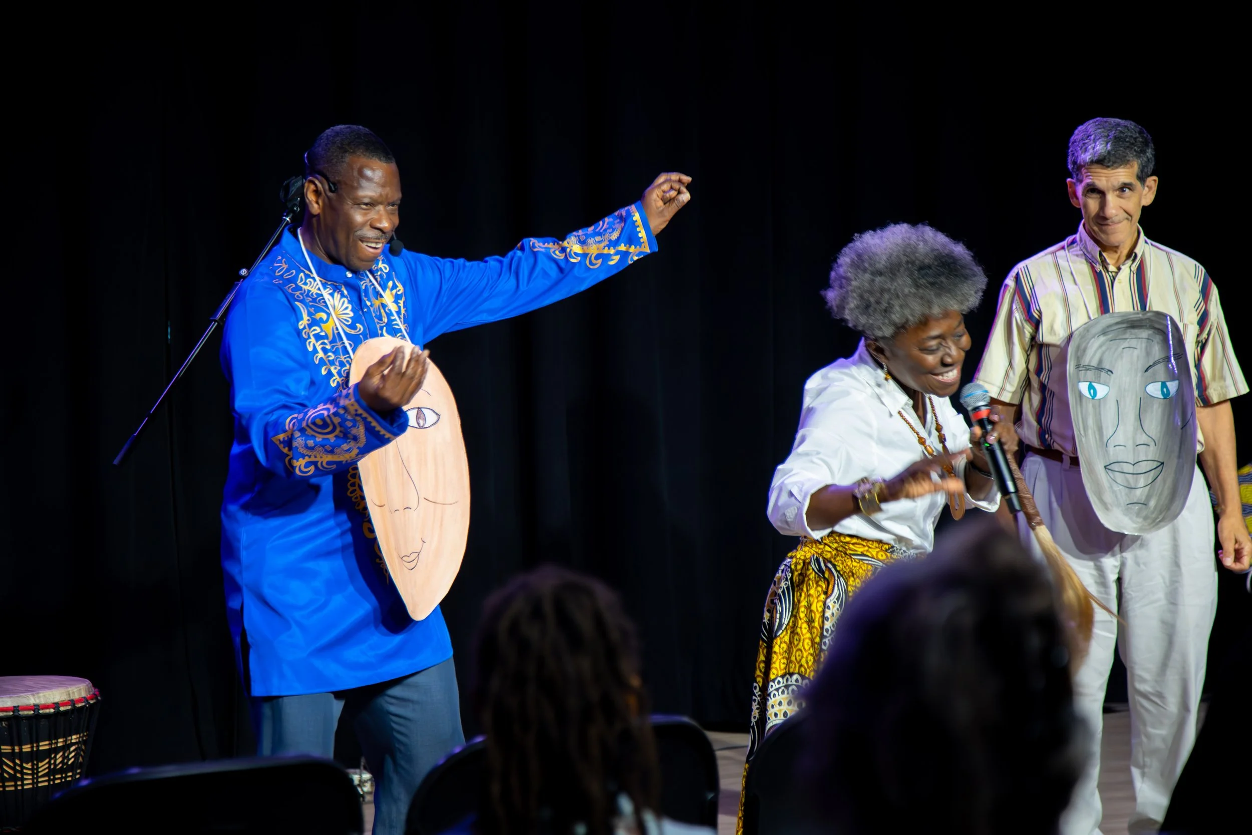 Three performers on stage, two men and one woman, holding masks and smiling, with a black curtain backdrop.