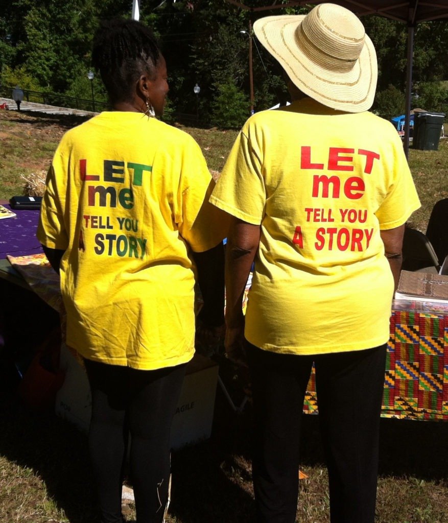 Two women wearing yellow T-shirts with the message "Let me tell you a story" printed on back, standing outdoors at a sunny event.
