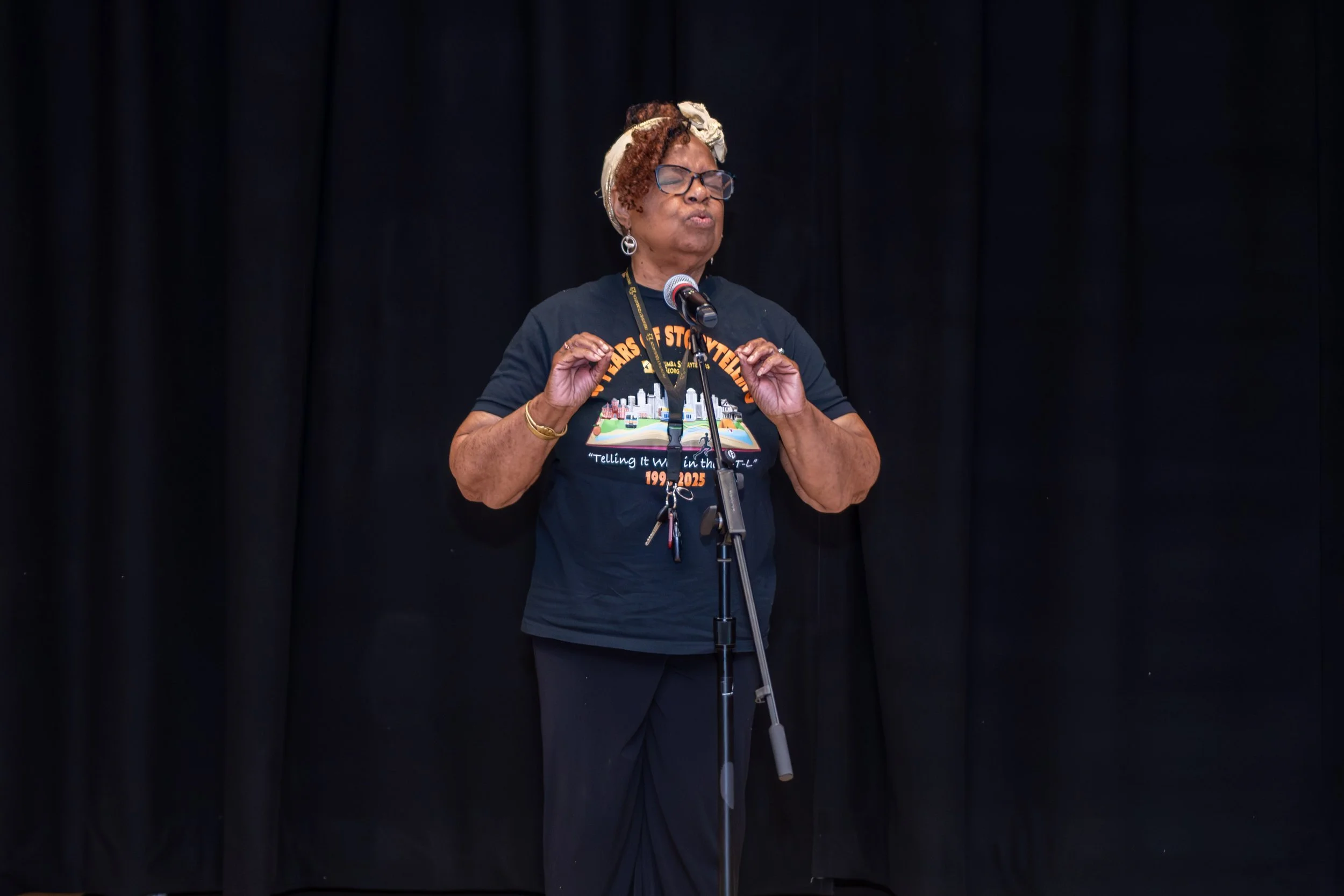 A woman with curly hair, glasses, and earrings speaking into a microphone on a stage with a black curtain background.