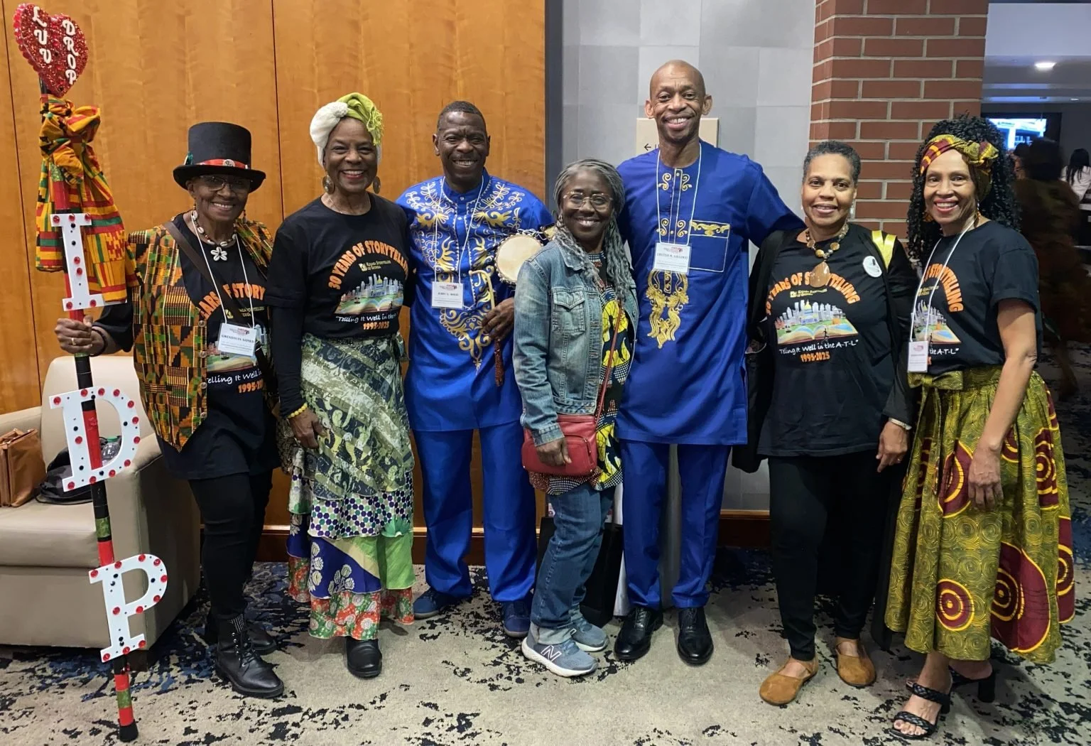 Group of eight people posing together indoors, some wearing traditional and colorful African attire, others in matching black T-shirts with a logo, and some holding decorative letters 'I', 'D', 'P'.