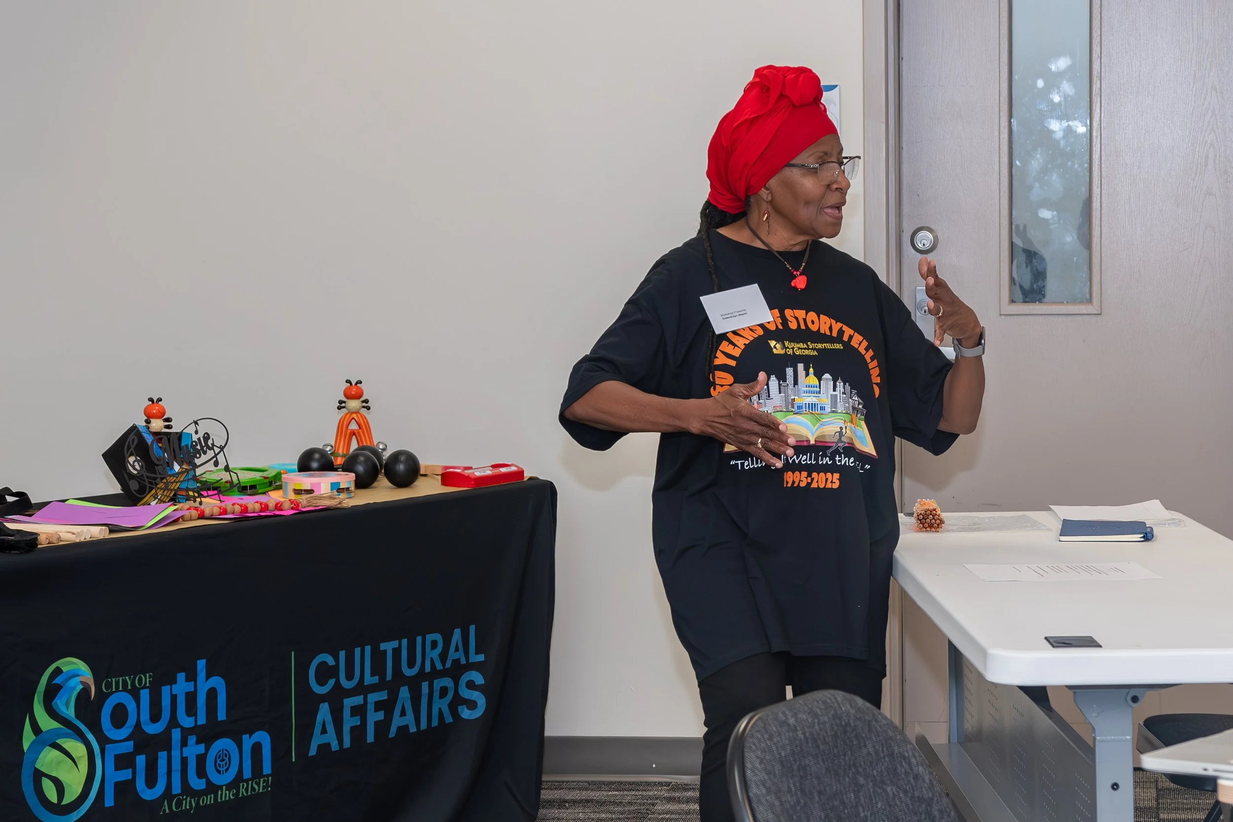 A woman wearing glasses and a red headwrap is speaking or gesturing while standing next to a table with various colorful objects and crafts at a cultural fair. The table has a black banner with the words 'City of South Fulton' and 'Cultural Affairs'.