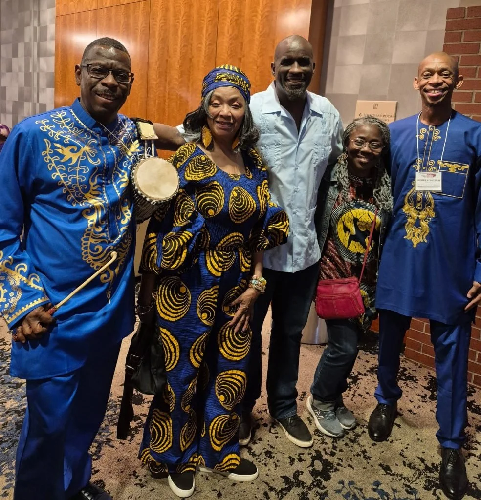 Group of five people standing together indoors, some wearing traditional African clothing, smiling for the photo