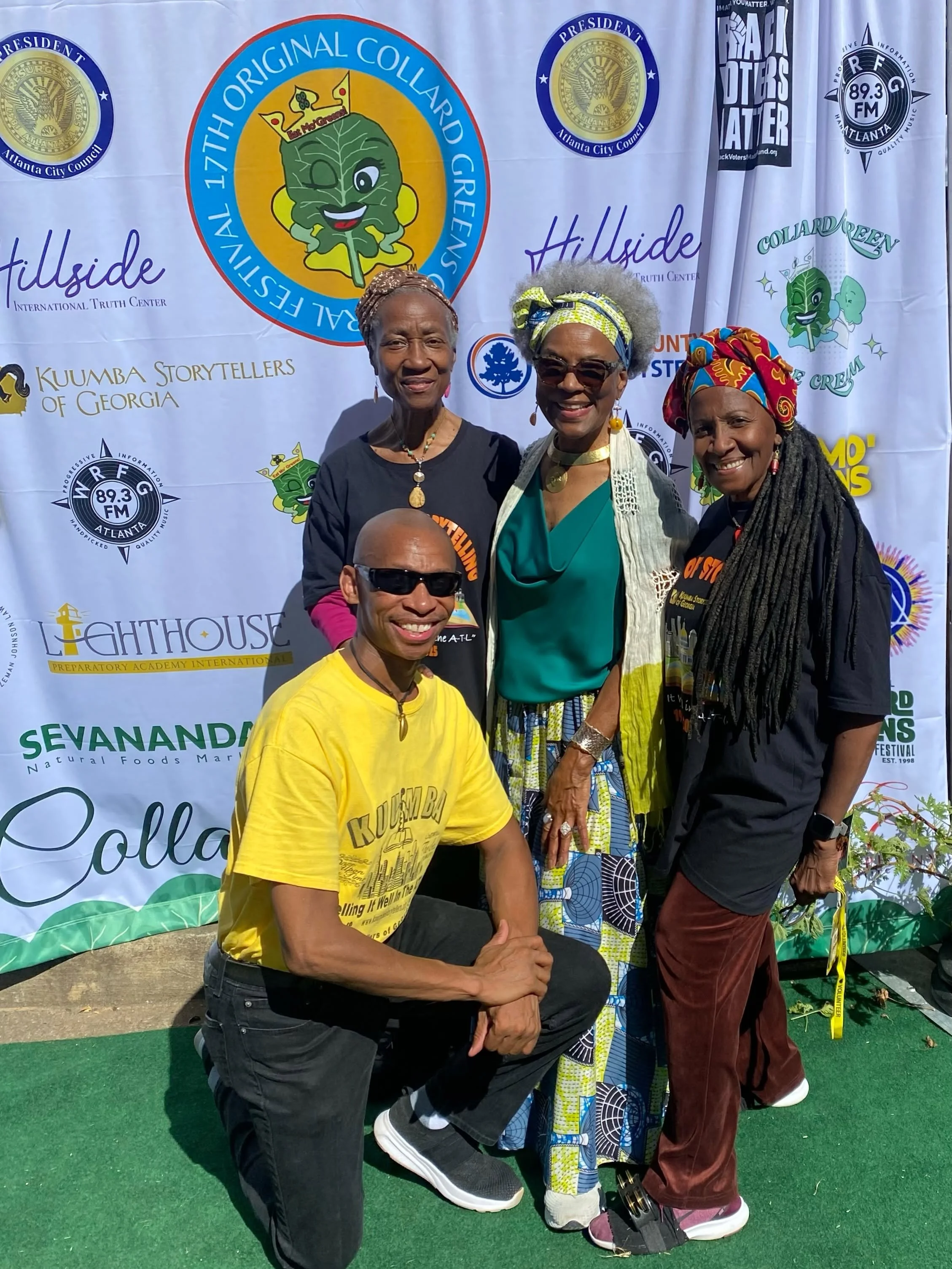 Four people, three women and one man, standing outdoors in front of a backdrop with various logos and text, smiling and posing for a photo. The backdrop includes logos for Atlanta City Council, local radio station, and other organizations.