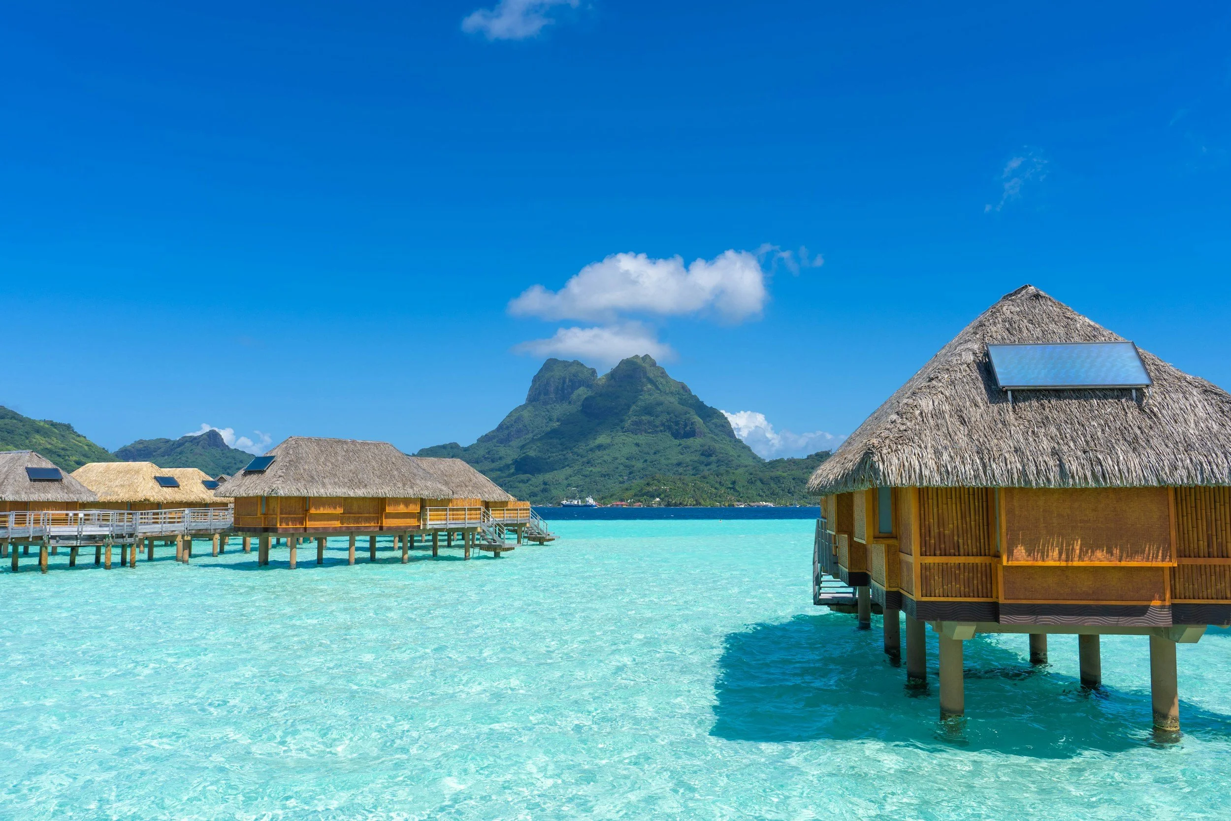 Overwater bungalows with thatched roofs and solar panels, turquoise water, and lush green mountains in the background under a blue sky.