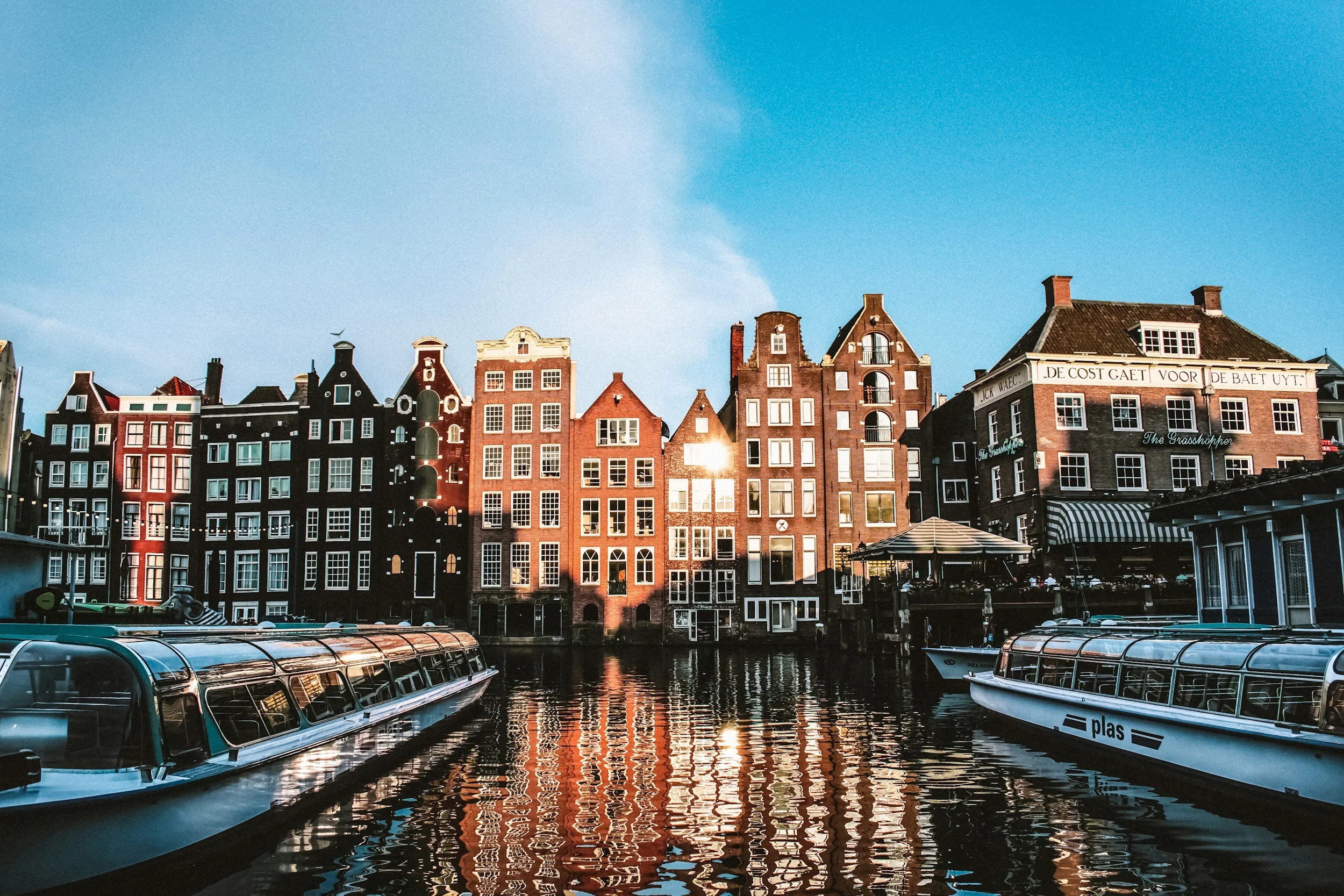 A row of traditional European buildings along a canal with boats docked in the water, under a clear blue sky during sunset.