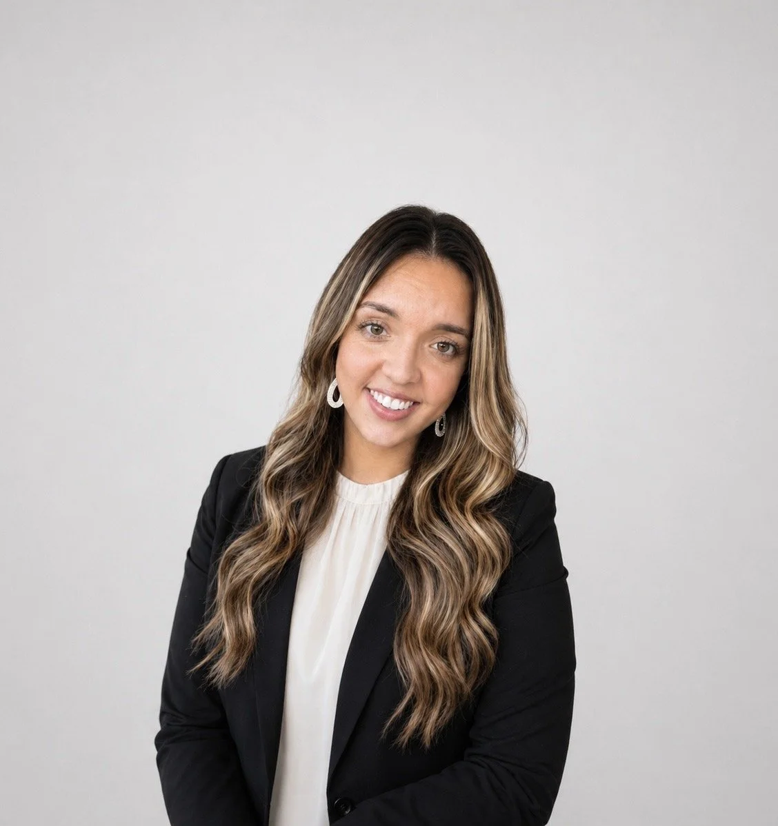 Portrait of a woman with long wavy hair wearing a black blazer and white blouse, smiling, against a plain light background.