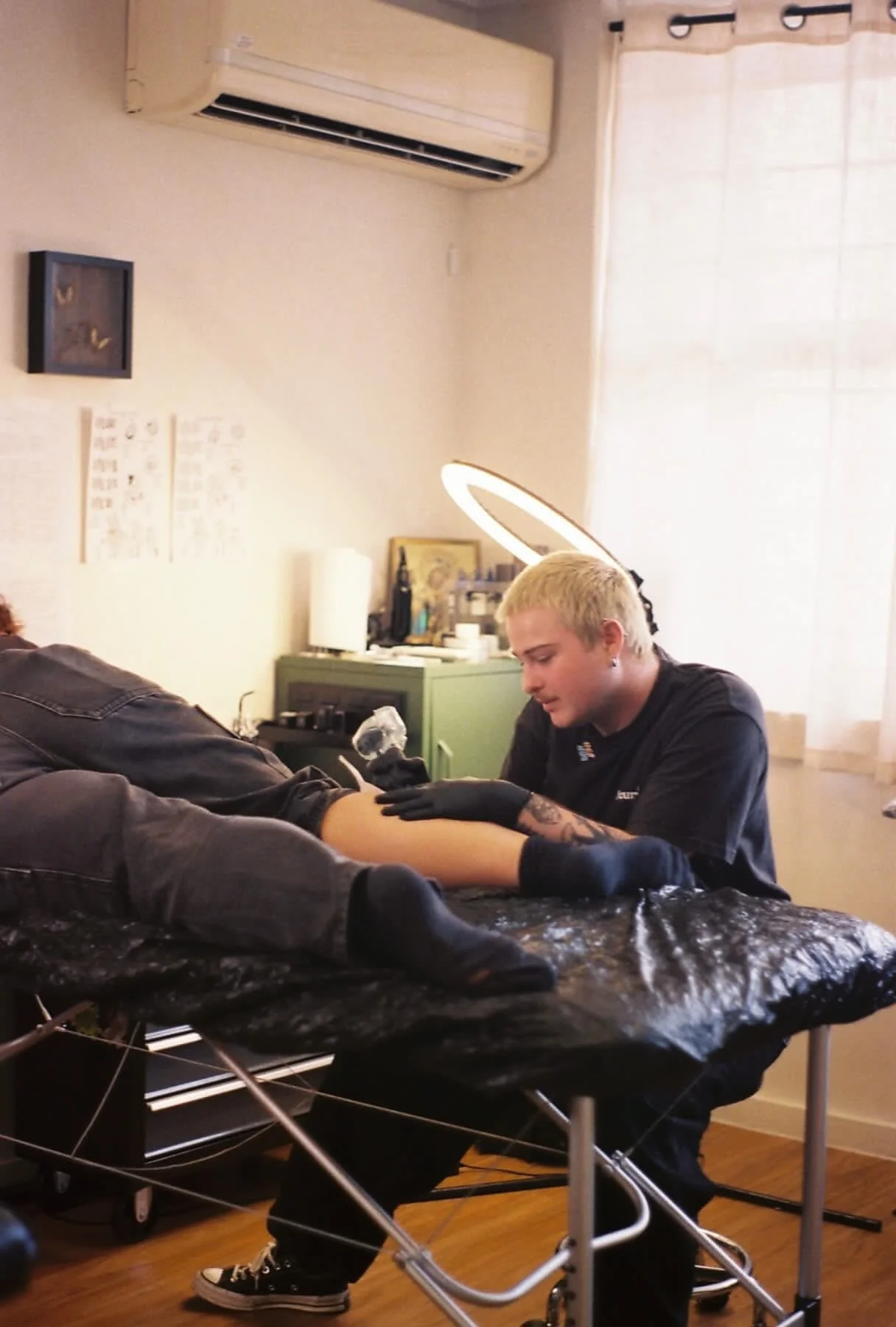 A tattoo artist with blonde hair and black gloves is tattooing a person's arm in a home setting, with a ring light illuminating the work area. The room has a green cabinet, framed pictures, and a window with curtains.